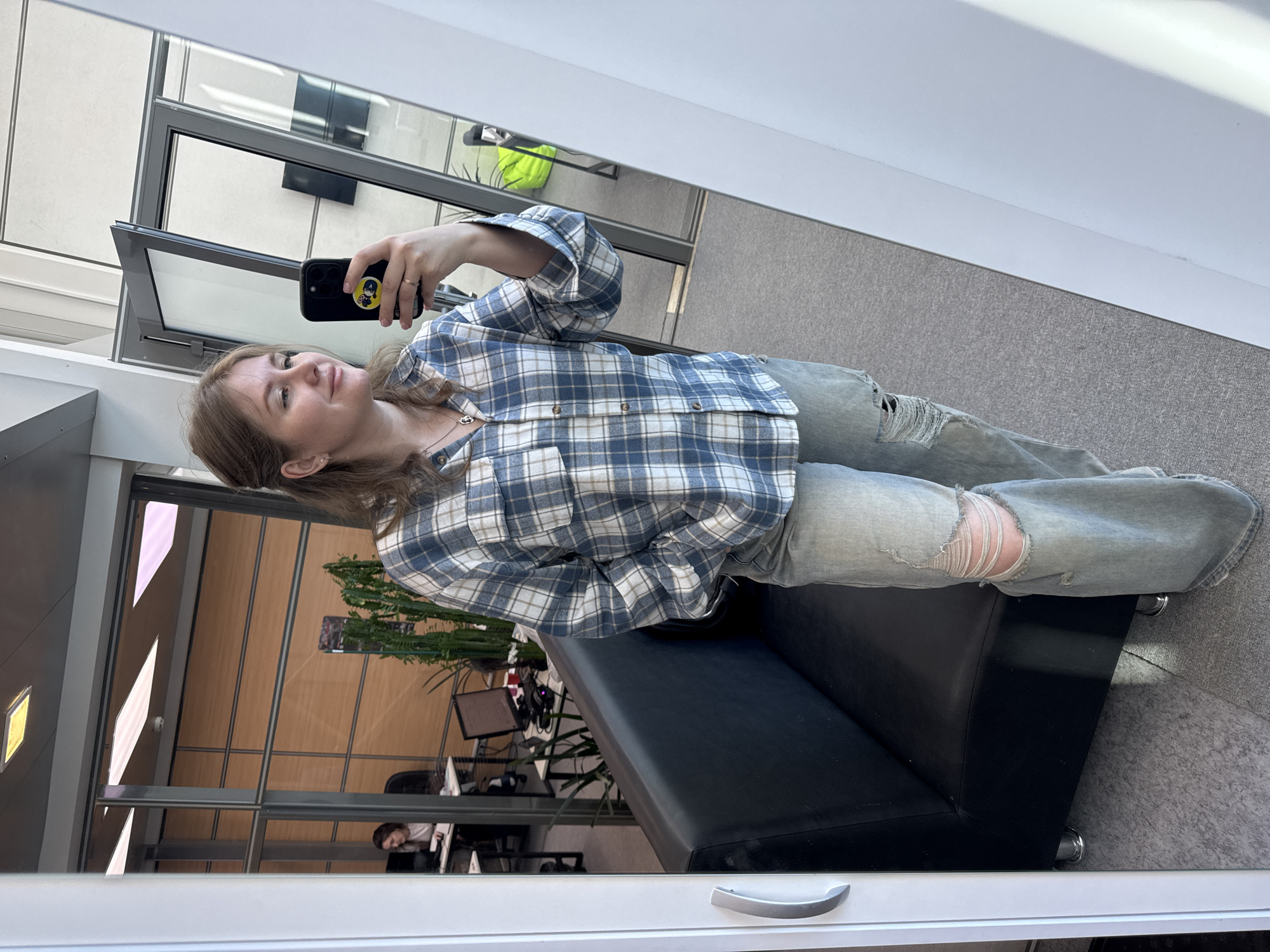 A person with fair skin and light brown hair is holding a smartphone and taking a selfie in what appears to be a mirrored or reflective surface, possibly a window. The individual is wearing a blue and white plaid button-down shirt and distressed light gray jeans with a tear at the knee. They are seated on a black leather-like bench or couch.
The background, as seen through the reflective surface, suggests an office or commercial building interior. Visible are cubicle-like workspaces with desks, computers, and office chairs. There are also indoor plants adding greenery to the space. The architectural style of the building includes large windows and metal framing. In the reflection, a person can be seen working at one of the desks in the office space.
The location cannot be identified based on the visual information provided in the image. There are no distinguishing landmarks or signs indicating a specific place within Perm, Russia.