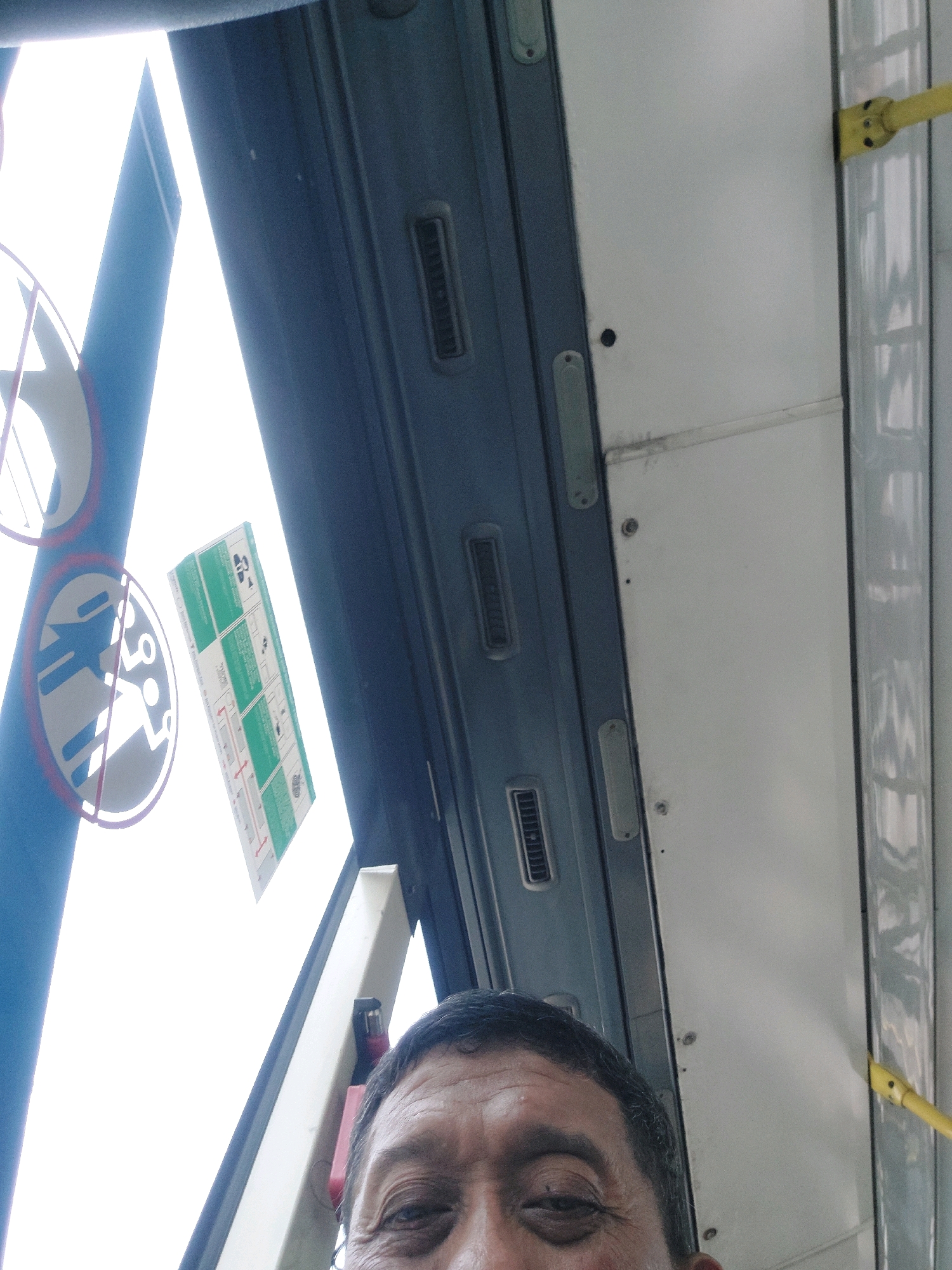 The image is a close-up, upward-angled selfie taken from inside a public transport vehicle, likely a bus, in East Jakarta, Indonesia. The perspective focuses on the upper part of a person's face, with their forehead, eyes, and the bridge of their nose prominently visible. The individual appears to be of East Asian descent. Behind the person, the interior of the vehicle is visible, characterized by a blue-colored panel on the left, a white panel on the right, and a light-colored ceiling. There are also some signs and warning decals affixed to the interior surfaces. One circular decal features a stylized silhouette of a person with a red diagonal line crossing through it, suggesting a prohibition. Another rectangular decal, with green and white elements, appears to be instructional or informational, possibly a safety or emergency guide. The yellow handrails, common in public transport, are also visible along the right side of the image. The lighting suggests an overcast day or an interior setting with diffused light coming from outside. The overall impression is candid and informal.