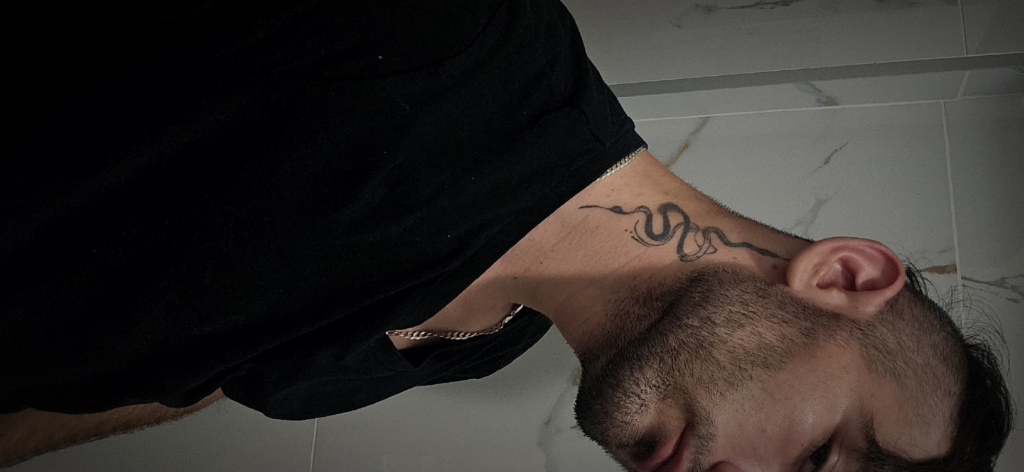 A person with short dark hair, a beard, and a snake tattoo on their neck is wearing a black shirt and a gold chain. They are in a room with white and grey tiled walls. No information is provided to determine if the picture was taken in Plovdiv, Bulgaria.