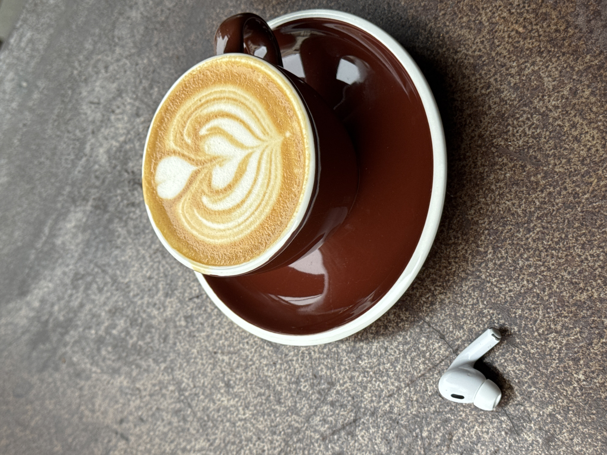 A close-up, top-down shot shows a brown ceramic coffee cup with a white rim, filled with a latte featuring latte art, resting on a matching saucer. To the lower right of the saucer is a single white wireless earbud. The background is a textured gray surface. There is no evidence to suggest the location of the city of Tel-Aviv, Israel.
