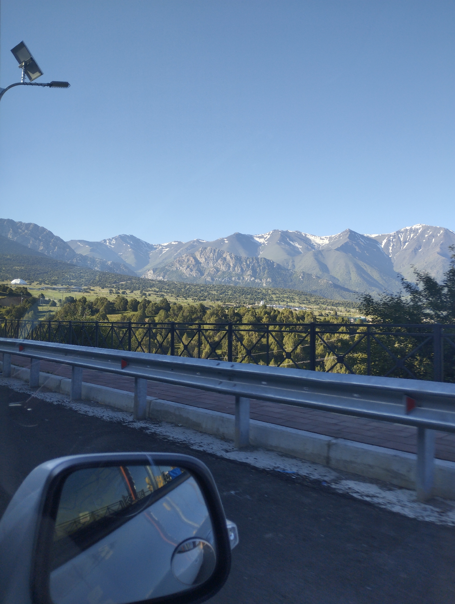 The image shows a roadside view with a mountainous landscape in the background. In the foreground, a car's side mirror reflects the road and surrounding scenery, including a metal guardrail and a pedestrian walkway. A solar-powered street lamp is visible on the left side of the image. The background features lush green hills dotted with trees, leading up to a range of snow-capped mountains under a clear blue sky. The overall scene suggests a scenic route outside a populated area. The image is not located in the city of Myk, Uzbekistan.