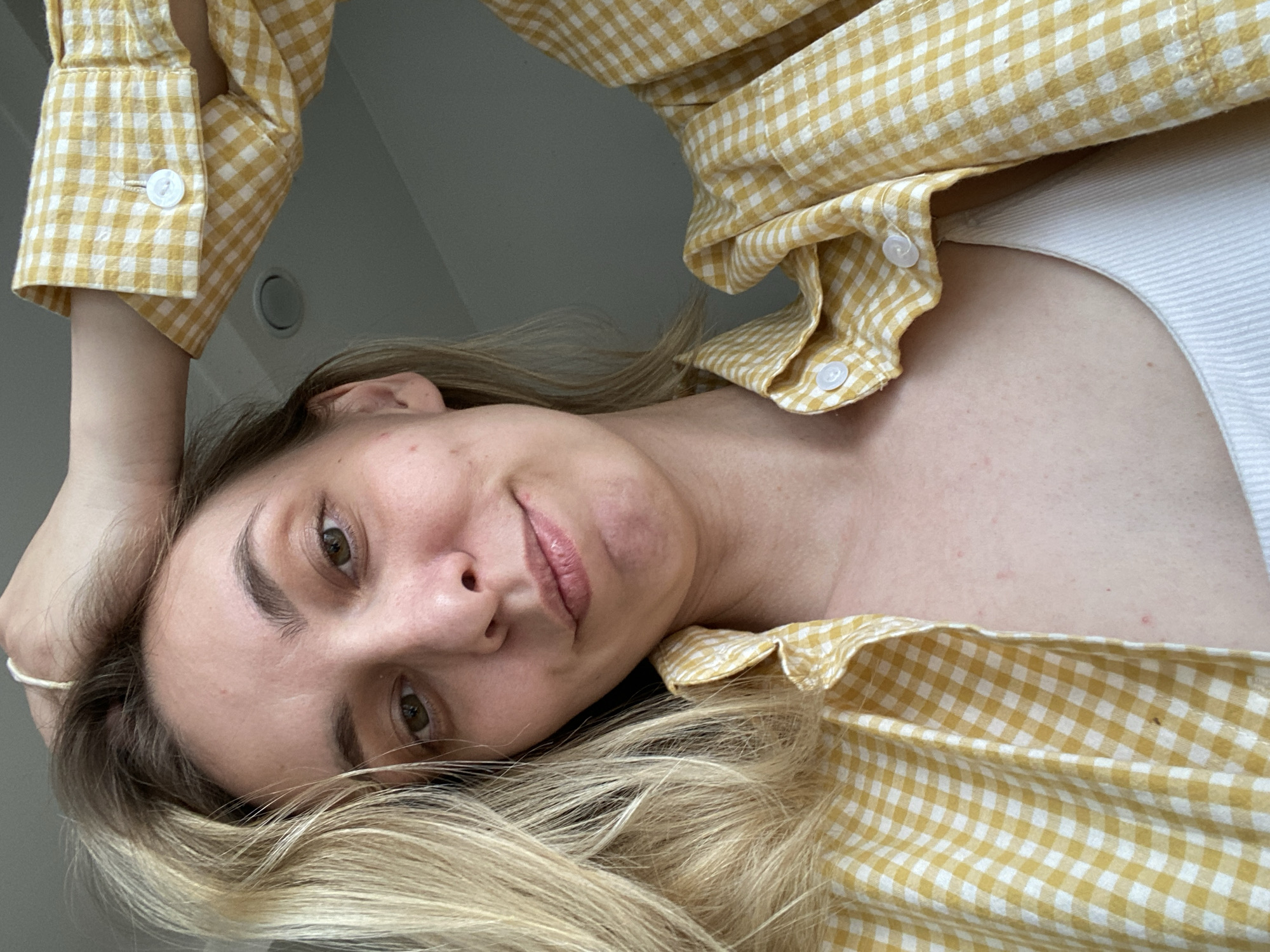 A woman with blonde hair is the primary subject of this photograph. She is wearing a yellow and white gingham-patterned button-down shirt and a white tank top. Her head is tilted, and her expression is contemplative. The background appears to be an interior space with a plain grey wall visible in the upper left corner. The location in the city of Chaikovsky, Russia is not determinable from the image.
