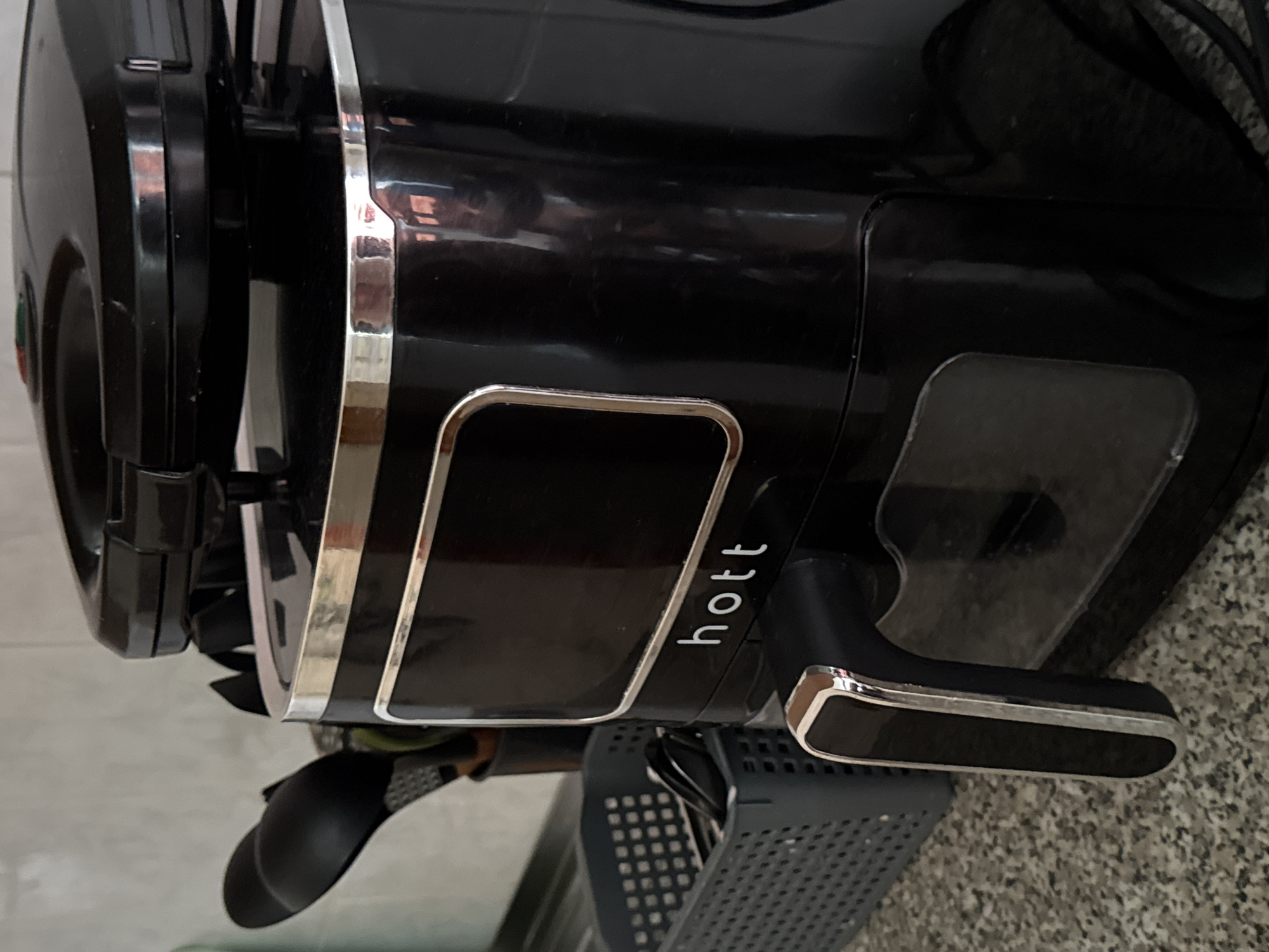 A black HOTT brand air fryer sits on a speckled gray countertop. The air fryer has a chrome trim around its display panel and handle. To the left of the air fryer is a grey plastic basket containing black utensils. In the background, a tiled wall and more kitchen utensils are visible. The scene is indoors, likely a kitchen, in Яромінскі сельскі Савет, Belarus.