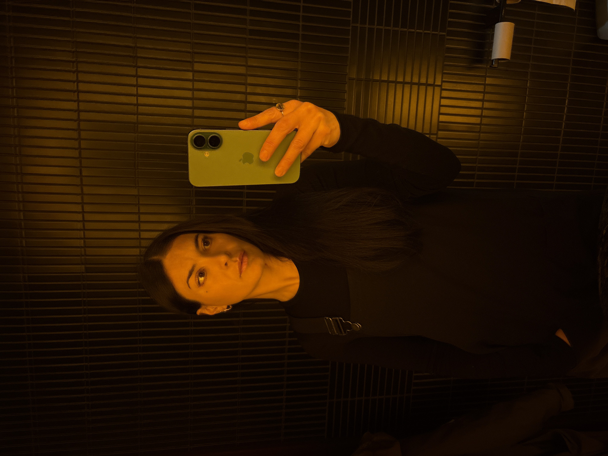 A woman is taking a selfie in a bathroom. She is holding a green iPhone in her right hand. She is wearing a black long-sleeved shirt and has long, dark hair. The bathroom has black tiled walls. A roll of toilet paper is visible in the upper right corner of the image. The bathroom appears to be dimly lit with an orange-yellow light. There are no clear indicators in the image to definitively place the bathroom in London, United Kingdom.