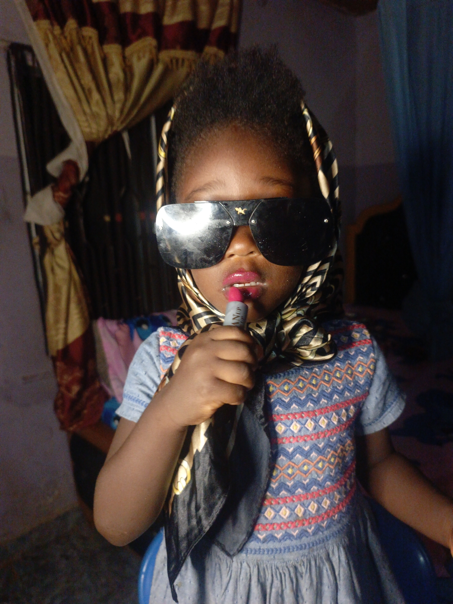A young child wearing sunglasses and a headscarf is applying lipstick. The child is wearing a blue dress with a patterned bodice. The background includes curtains and other household items. This photo was likely taken indoors in Akwanga, Nigeria.