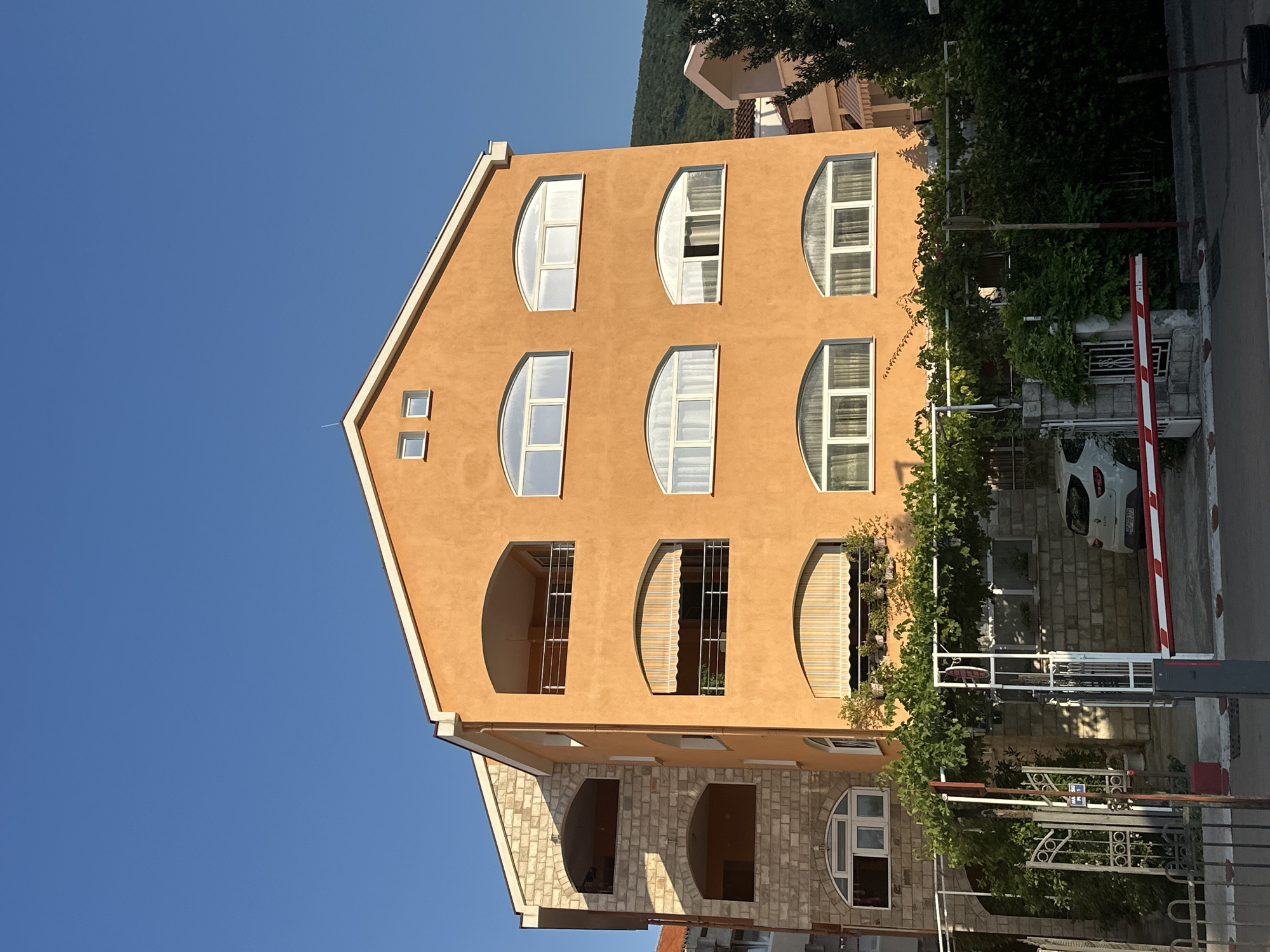 The image captures a multi-story building with an orange facade, typical of the architecture found in Budva, Montenegro. The building features several arched windows and balconies. To the left, an adjacent structure is partially visible, constructed with stone. Lush green foliage, including what appears to be a grapevine, covers parts of the building's exterior and a metal pergola in front of it. A white car is parked near the entrance, which is secured by a white and red barrier. The scene is set against a clear blue sky, and a treed hillside is visible in the background. The lighting suggests it is daytime, likely late afternoon due to the warm tones.