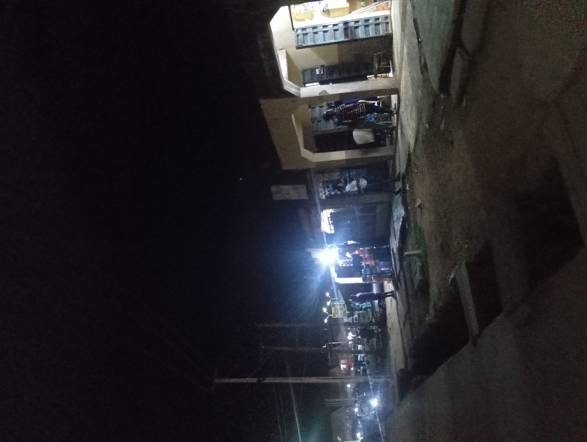 An exterior, low-angle, night-time shot captures a street scene in an urban environment. The foreground shows a concrete sidewalk and a dark, open drain. Further back, several businesses with open doorways and illuminated interiors line the street. Bright lights, possibly from streetlights or shop signs, create a strong contrast with the dark sky. A few people are visible standing or walking in front of the businesses. A utility pole with wires is prominent on the left side of the image, and a vehicle with its headlights on is present in the middle ground. The overall impression is of a bustling, albeit dimly lit, commercial area at night.