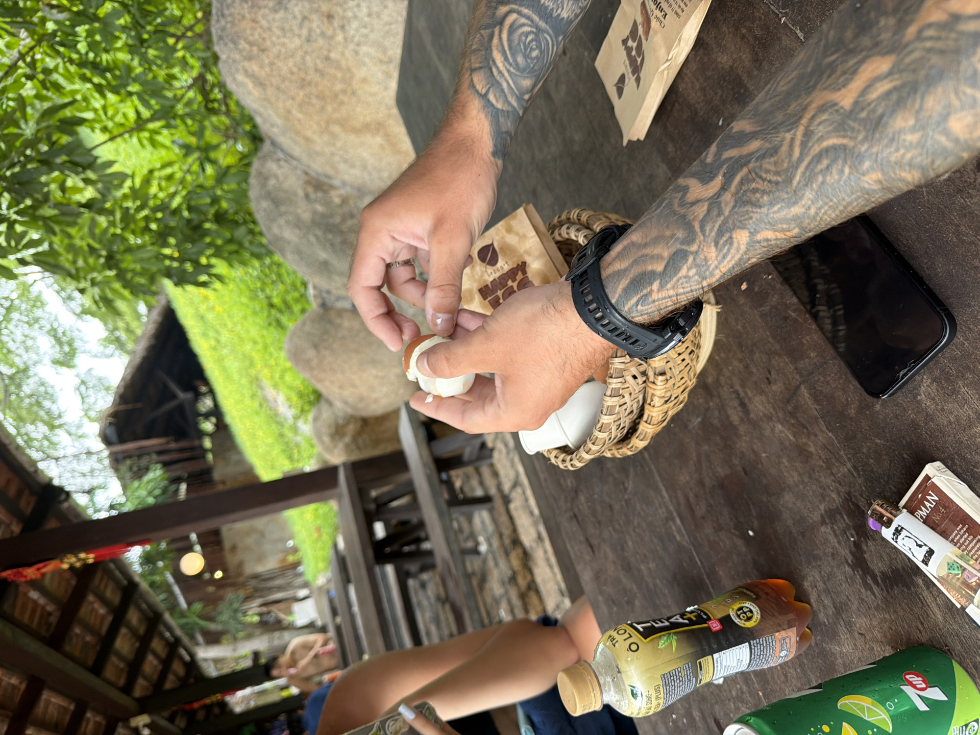 An overhead, medium shot captures a close-up view of a wooden table outdoors, where a person's hands are shown peeling a hard-boiled egg. The background reveals a rustic, open-air establishment, likely a cafe or restaurant, with thatched roofing and wooden structures, set against a backdrop of lush green foliage and large, rounded rocks. The table itself is laden with various items including a bottle of iced tea, a can of 7UP, a pack of cigarettes with a lighter, a smartphone, and a woven basket containing a small cup. The person's arms are prominently adorned with intricate black and grey tattoos. The scene suggests a casual outdoor dining or relaxation setting.