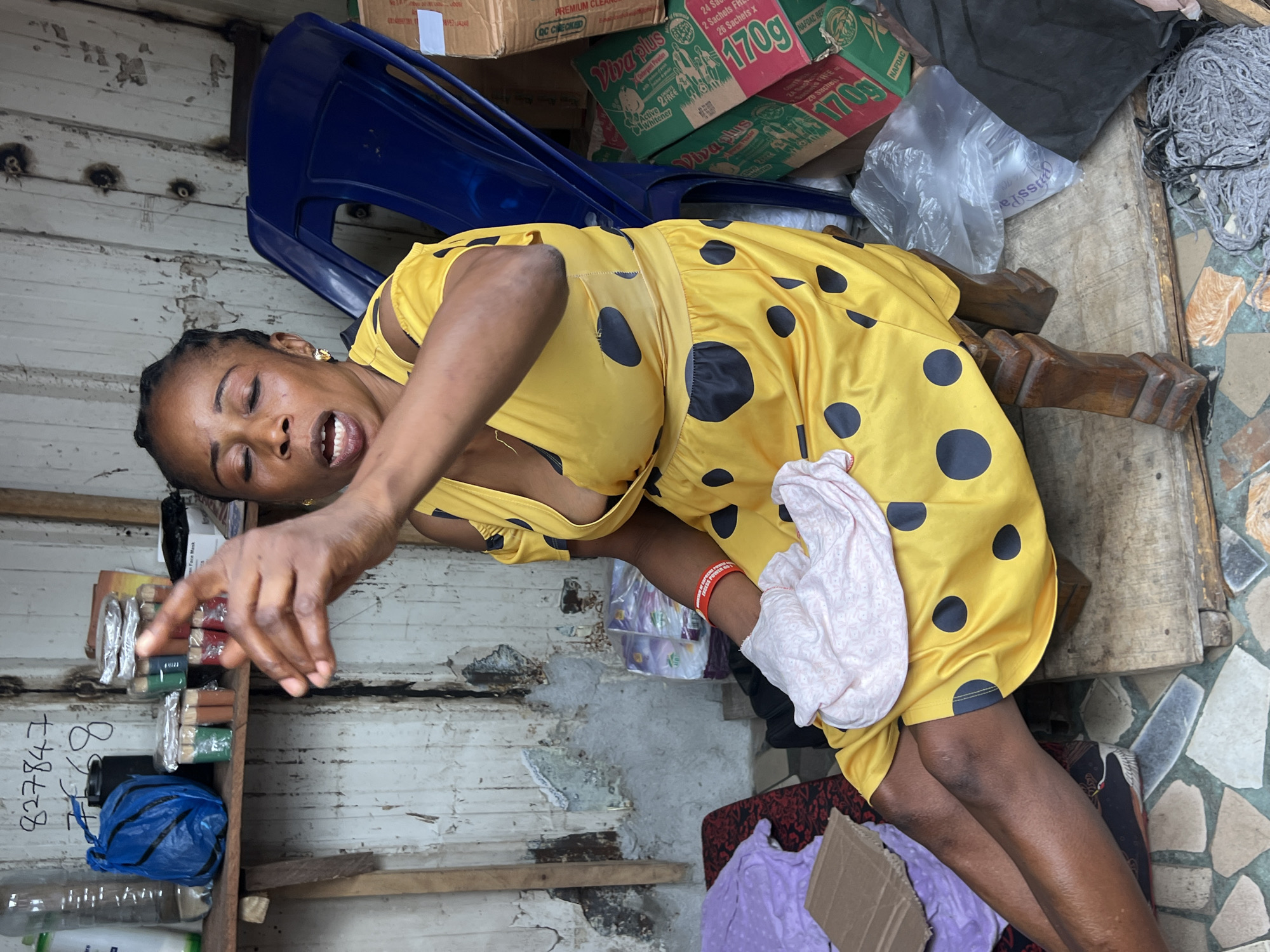 An African woman in a yellow polka dot dress with a plunging neckline is sitting on a wooden stool in Nkpor, Nigeria. Her eyes are closed, and her mouth is open as if she is speaking or singing. Her right arm is raised, and her index finger is pointing forward. She is holding a light pink piece of fabric in her lap. The background consists of a weathered, white corrugated metal wall and a blue plastic chair. Several boxes are stacked behind the chair, one of which has green and red branding that reads "Viva Plus". A shelf above the woman holds several bottles and small containers. The floor is tiled with a mosaic of irregular stone-like pieces. To the right of the stool, there is a black plastic bag and a bundle of gray rope. The scene appears to be an outdoor or semi-outdoor setting, possibly a market stall or a makeshift shop.