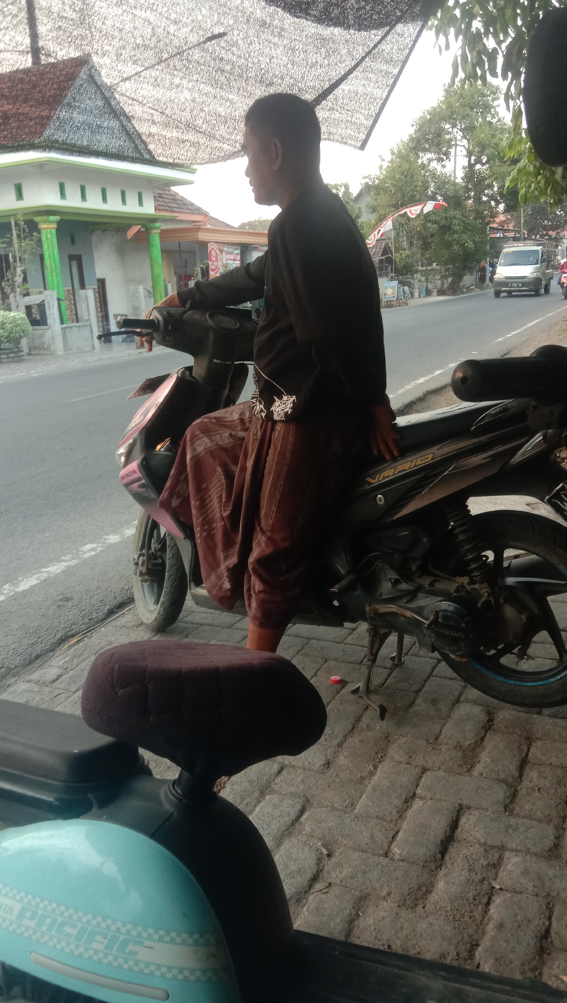 A man wearing a black long-sleeved shirt and a patterned brown sarong sits on a black Honda Vario motorcycle parked on a paved sidewalk next to a road. The motorcycle has a pink sticker on its front fairing that says "HONDA". To the left of the motorcycle, another motorcycle with a light blue front fender is partially visible. The background shows a street with trees and buildings, including a house with green columns and a red-tiled roof. The sky is overcast. The scene appears to be in a tropical, urban or semi-urban environment, likely Karang Anyar, Indonesia, given the architecture and vegetation.