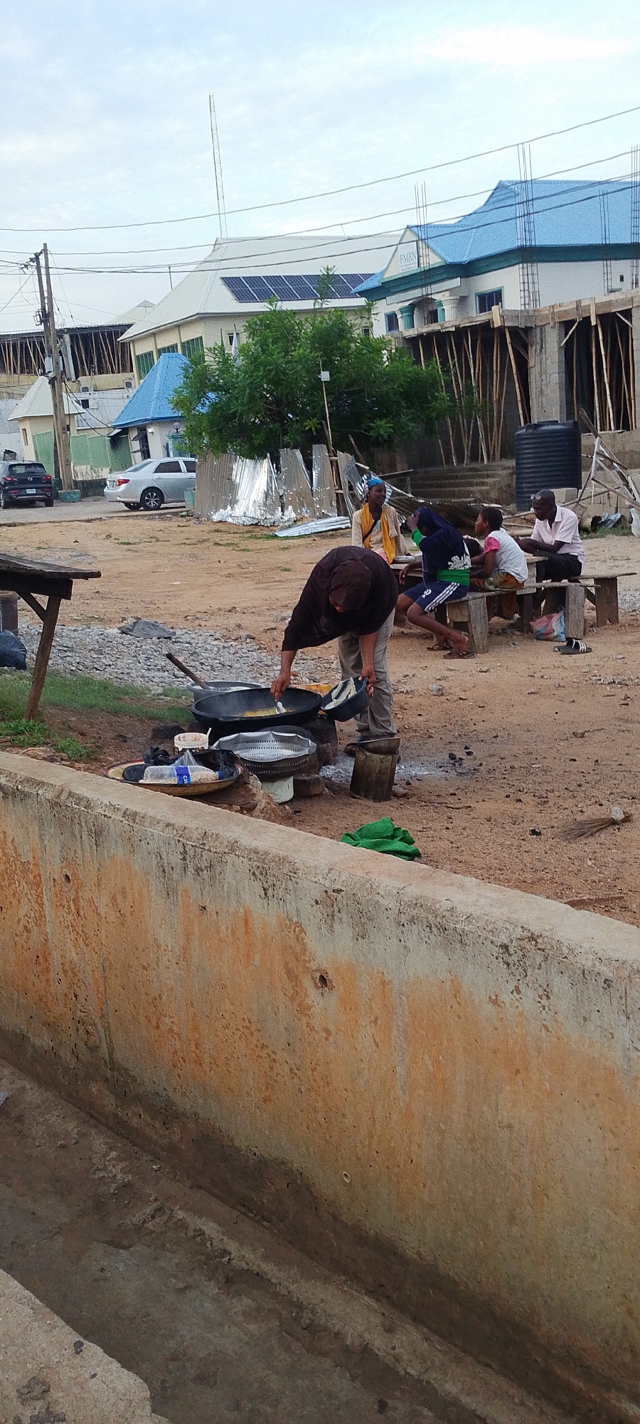 The image shows an outdoor scene in Lokoja, Nigeria. In the foreground, a low concrete wall runs along the bottom of the frame. Behind it, a person is cooking food in a large wok placed on a stove-like structure made of rocks. The person is bent over the wok, appearing to stir or add ingredients. There are other cooking utensils and containers nearby.
In the background, a group of four people are seated on wooden benches, seemingly observing the cooking process. Behind them is a makeshift structure of corrugated metal and an unfinished building under construction, with wooden supports. The sky is overcast.
Two cars are visible parked on the road. Overhead power lines are visible along the top of the image.
