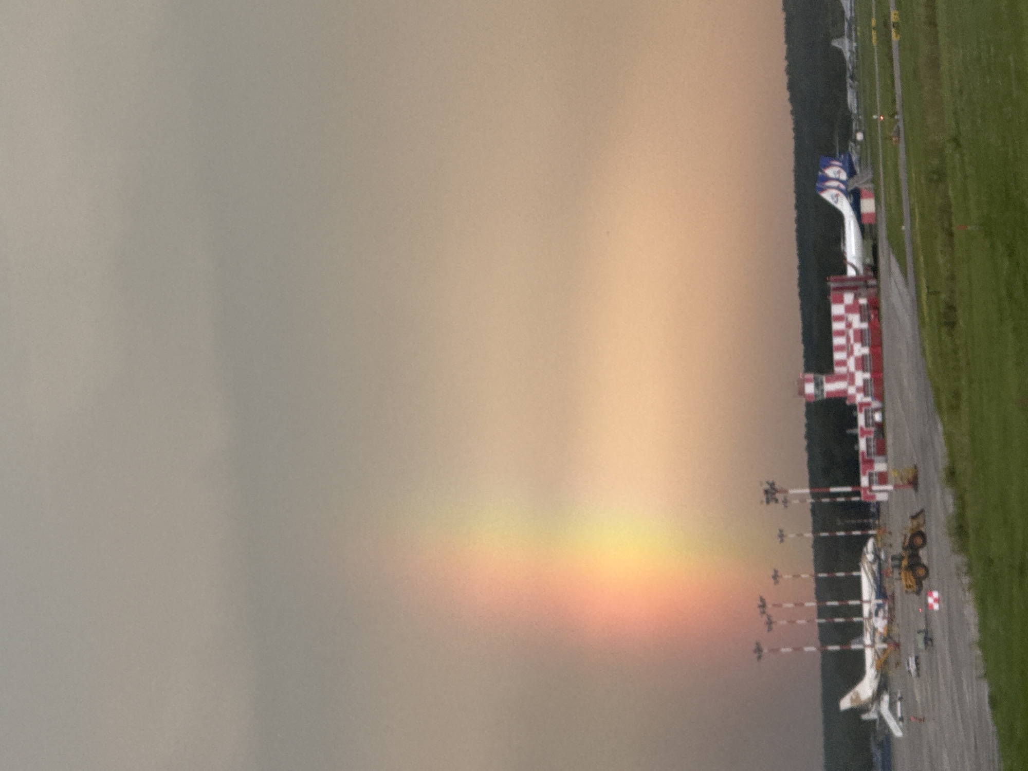 This image depicts an airport tarmac at dusk, with a rainbow visible in the overcast sky. The scene is oriented vertically, suggesting the original photo was taken in landscape and rotated. On the tarmac, several small aircraft are parked, some with service equipment around them. A red and white checkered tower stands prominently, likely an air traffic control or airport building. To the right, a grassy area runs alongside the tarmac, and a few vehicles, including what appears to be a yellow loader, are present. The sky is a gradient of muted colors, transitioning from a dusky grey on the left to warmer, orange hues on the right, where the rainbow arc is faintly visible, showing bands of yellow and red. The overall impression is of a quiet moment at the airport as daylight fades, with the rainbow adding a touch of ethereal beauty to the otherwise functional setting. The specific location is Yekaterinburg, Russia. No people are clearly visible, and there is no discernible text in the image.