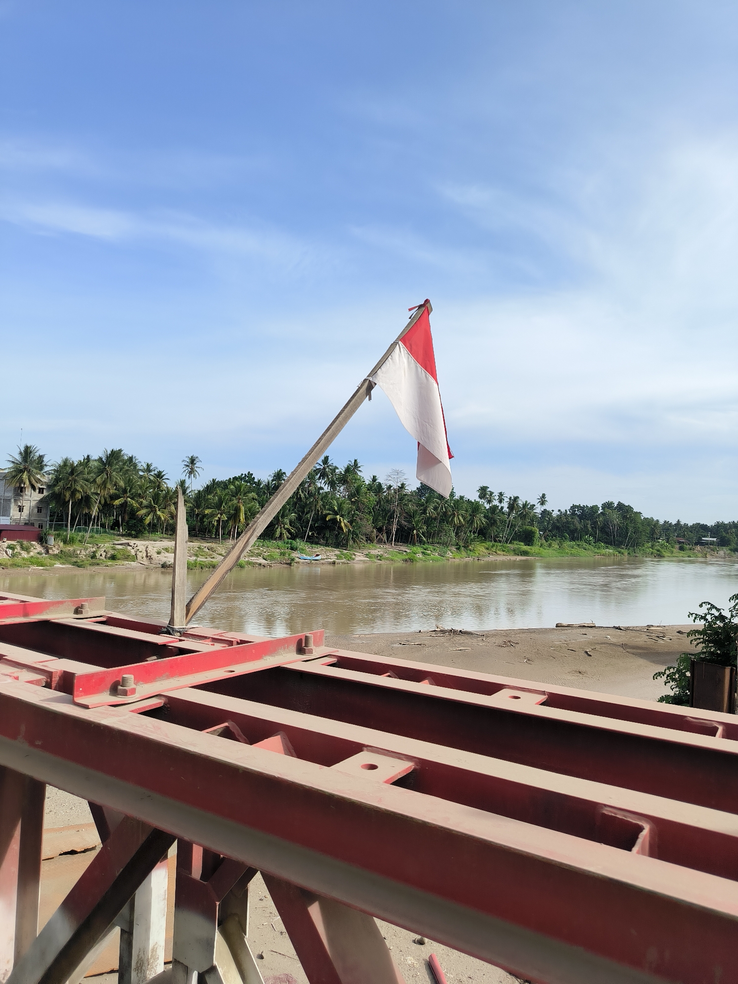 The image captures a scene along the Mahakam River in Tingkeummanyang, Indonesia. In the foreground, a red metal structure, possibly part of a bridge or a ferry, dominates the lower half of the frame. A weathered wooden pole extends diagonally upwards from this structure, bearing the Indonesian flag, which is split horizontally into red and white. The river, a muddy brown, occupies the middle ground, with a sandy embankment visible on its far side. This embankment is lined with a dense tropical forest, characterized by numerous palm trees and other lush vegetation. A few buildings, including a light-colored multi-story structure, are partially visible amongst the trees on the left bank. The sky above is a bright blue with scattered white clouds. The overall impression is one of a rural riverside landscape, with man-made structures interspersed with natural elements under a clear sky.