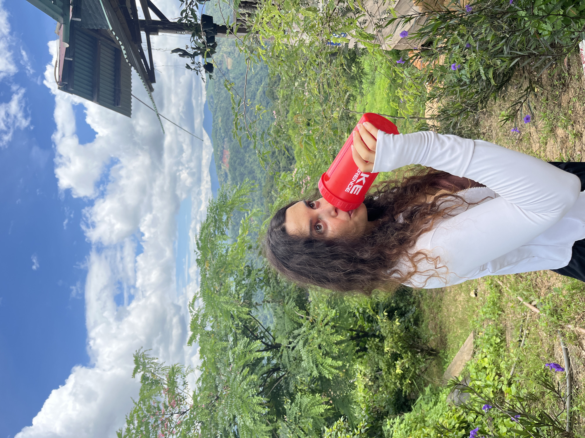 A woman with long, dark, curly hair is shown from the chest up, drinking from a red shaker bottle. She is wearing a white long-sleeved shirt and black leggings. The background is a lush, green mountainous landscape under a blue sky with white clouds. A rustic wooden structure with a corrugated metal roof is visible to the right. The location is Sơn Điền, Vietnam.