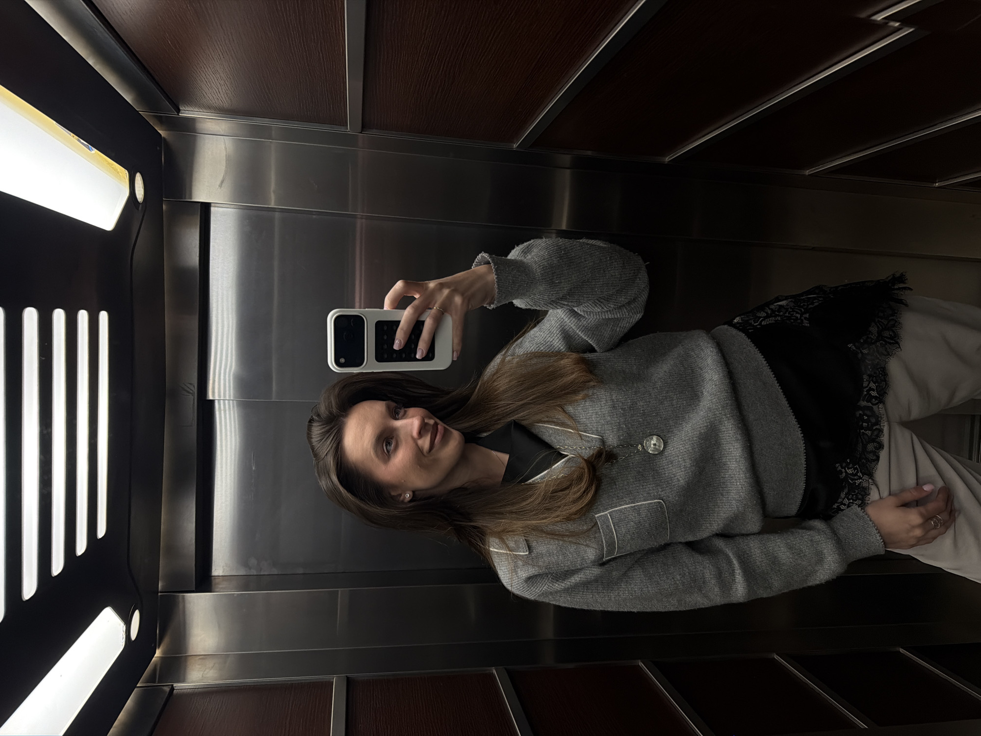 A woman is taking a selfie in an elevator with brown wood and silver metal paneling. The woman is facing left, wearing a gray cardigan with white trim, a black top with lace detailing, and light-colored pants. She is holding a white smartphone in her right hand. The elevator has vertical light panels on the left side. The location is not identifiable as Budva, Montenegro from the image.