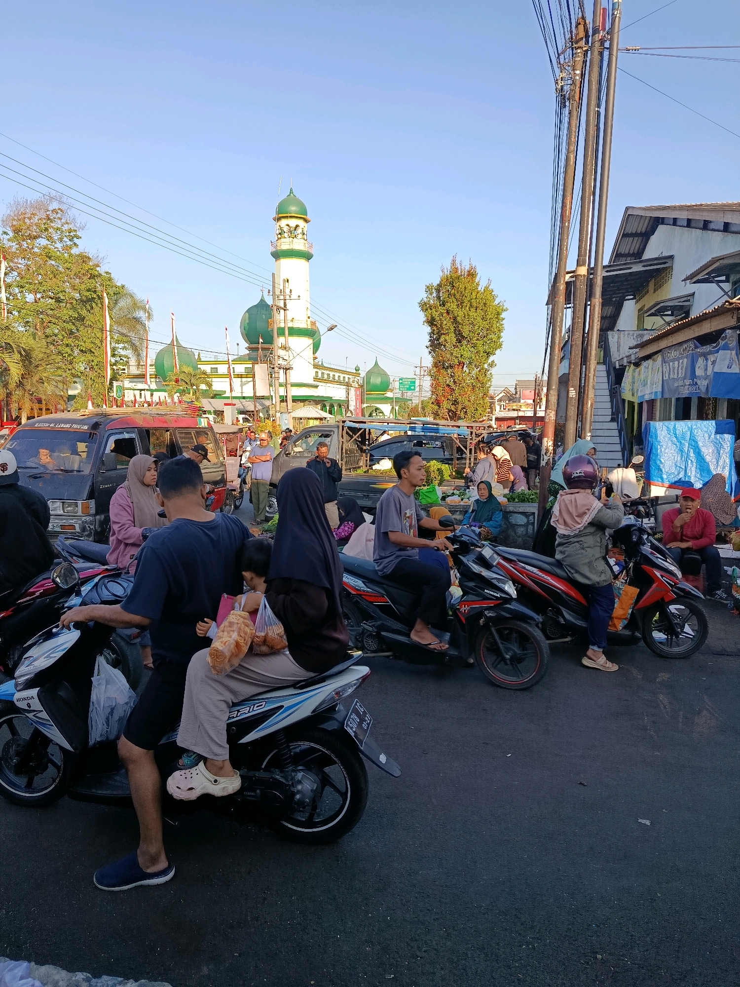 In Bululawang, Indonesia, on a sunny day, a bustling scene unfolds near a mosque. A family of three is seated on a motorcycle in the foreground, with the father driving, the mother holding a plastic bag of food, and a child in her lap. The father wears a dark blue shirt and shorts, while the mother is in a gray hijab and a long-sleeved pink shirt. They are all wearing casual footwear. Behind them, other people on motorcycles and in vehicles navigate the street. In the background, the distinctive green domes and white minaret of the mosque stand out against the clear blue sky. The area appears to be a marketplace, with stalls and vendors visible, adding to the lively atmosphere. The overall impression is one of everyday life in a lively Indonesian town.