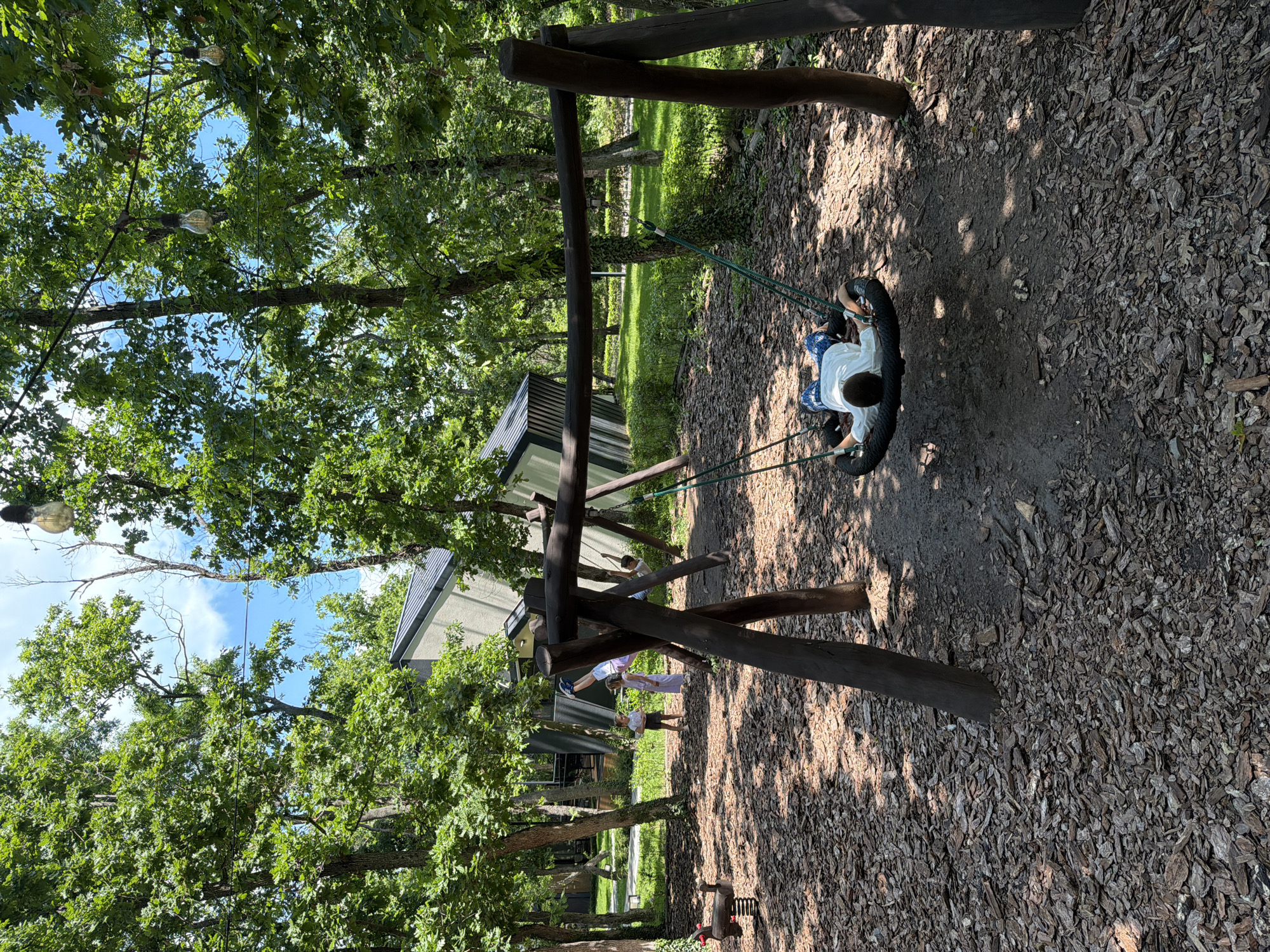 The image shows a children's playground located in a park. A child wearing a white shirt and blue patterned pants is sitting on a tire swing. The swing is suspended from a wooden frame. The ground is covered in wood chips. Other children and an adult are visible in the background near a building. The area is surrounded by trees, and the sky is visible through the leaves. A lightbulb hangs from a cable strung between trees. The specific location in Burgas, Bulgaria, cannot be determined from the image.
