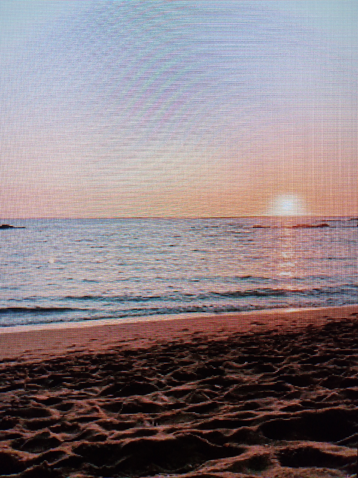 The image depicts a beach scene at sunset. The sky is a gradient of colors, transitioning from light blue at the top to orange near the horizon. The sun is setting over the ocean, casting a bright glow. The ocean is a mix of blues and oranges, reflecting the colors of the sky. Gentle waves are breaking on the shore. The beach is sandy, with footprints visible in the foreground. The location is unknown.