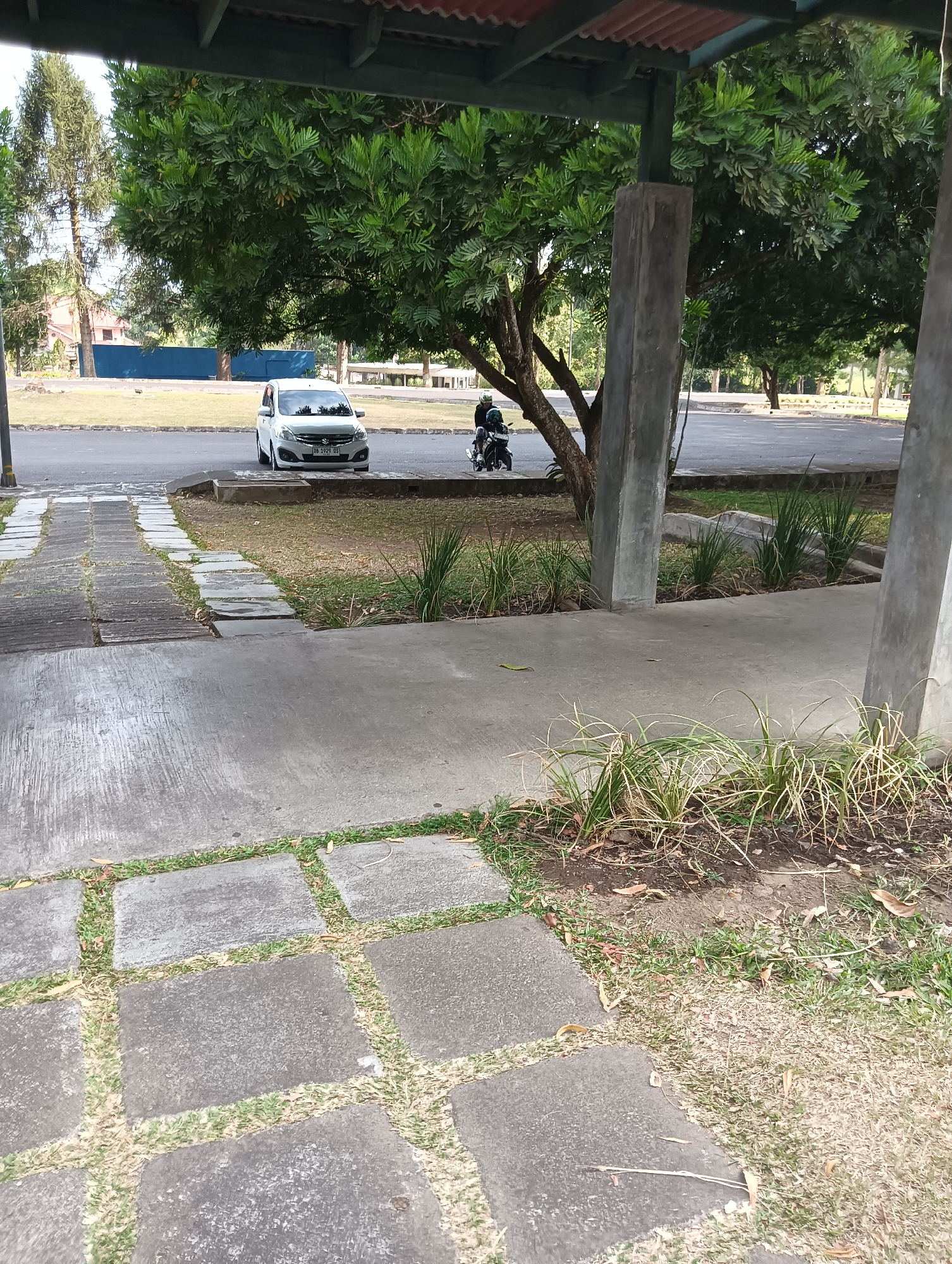 The image depicts an outdoor scene with a paved pathway in the foreground, leading towards a road and a grassy area. A white car is parked on the right side of the road, and a motorcycle with a rider is visible on the road as well. Large trees with lush green foliage are present throughout the scene, some of which are supported by concrete pillars. In the background, there are buildings and more trees, suggesting a park-like or institutional setting. The overall atmosphere is bright and appears to be daytime. The ground is a mixture of concrete slabs and grass.