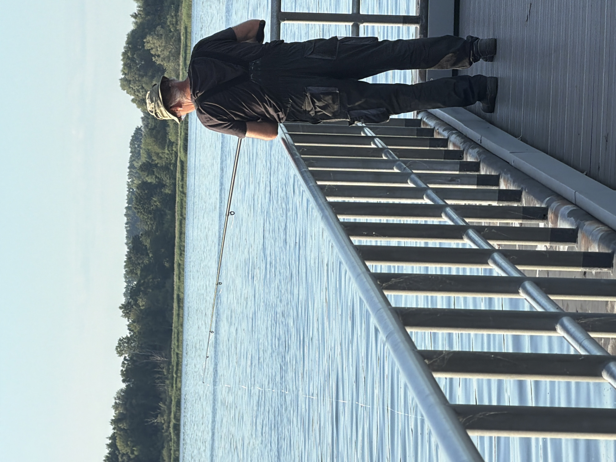 An adult male is fishing from a pier on a body of water. He is wearing a black t-shirt, black overalls with multiple pockets, and a camouflage hat. He holds a fishing rod, with the line extending into the water. The pier has a metal railing and a wooden surface. In the background, there is a line of trees and a clear sky. The scene is set in Ribnitz-Damgarten, Germany.