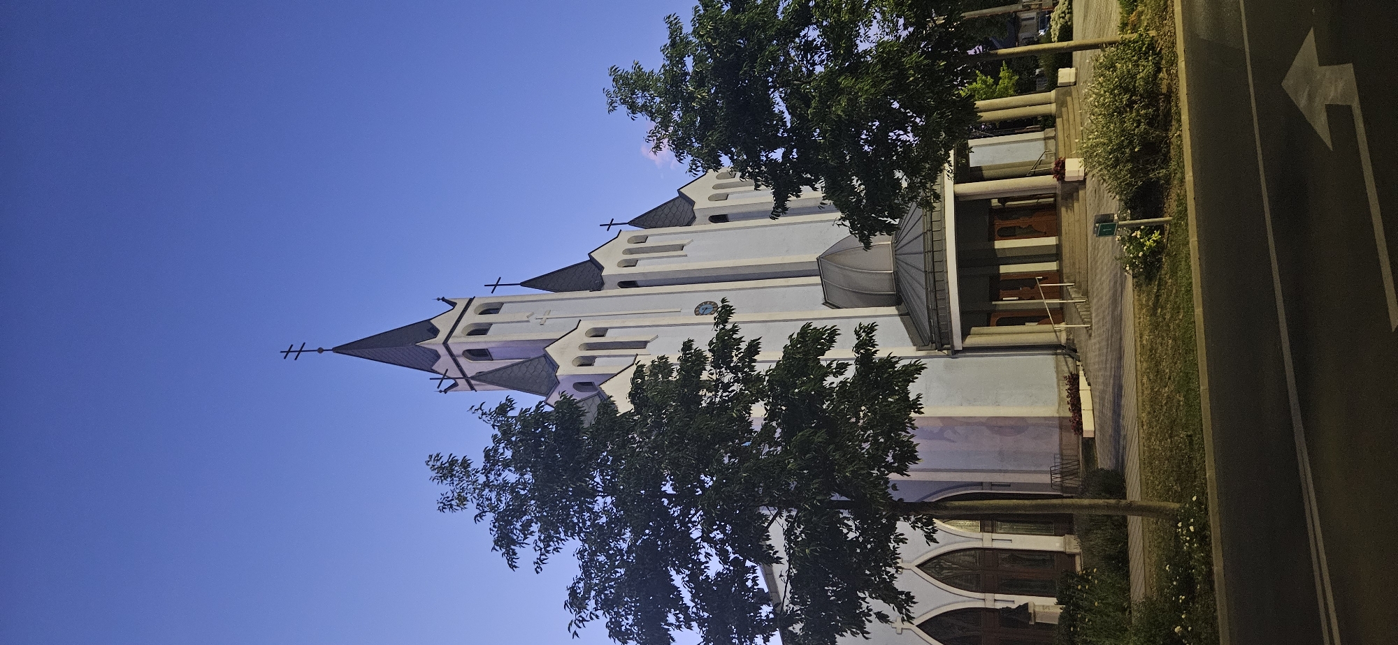 The image shows the exterior of the Hévíz Catholic Church in Hévíz, Hungary, at dusk. The church is a white building with a tall, dark steeple topped with a cross. There are several smaller spires and crosses adorning the roofline. Large trees with dark green foliage flank the front of the church, with one tree prominently positioned to the left and another to the right, partially obscuring the building. The sky above is a deep, clear blue, characteristic of twilight. In front of the church is an entrance with a covered portico supported by columns, leading to double wooden doors. There are also steps leading up to the entrance. The surrounding area appears to be landscaped with grass, bushes, and some flowering plants. A road with a white directional arrow painted on it is visible in the foreground. The lighting suggests it is not daytime, with some artificial illumination possibly present on the church. No people are visible in the image.