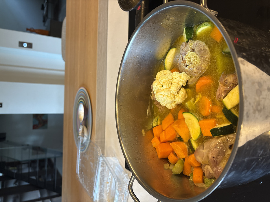 A large metal cooking pot, approximately two-thirds full of a yellow broth containing various ingredients, is centered on a black induction stovetop in a kitchen in Raanana, Israel. The pot's contents include numerous diced orange carrots, slices of green zucchini, several pieces of light-colored meat (possibly oxtail or similar cuts), and at least two large florets of cauliflower.
In the mid-ground, a light-colored wooden kitchen island or counter extends horizontally. On this counter, to the left of the pot, an open clear plastic food container with its lid resting nearby is visible. To the right of the container, a circular multi-colored striped plate holds two small, metallic-colored bowls or serving spoons.
The background shows a blurred interior space, indicative of an open-plan living area. Elements discernible include a darker vertical structure, possibly a staircase railing, on the left, and white walls with what appears to be abstract artwork mounted. Light switches or wall outlets are also visible on the white walls. The overall environment suggests a modern residential kitchen. No people are visible.