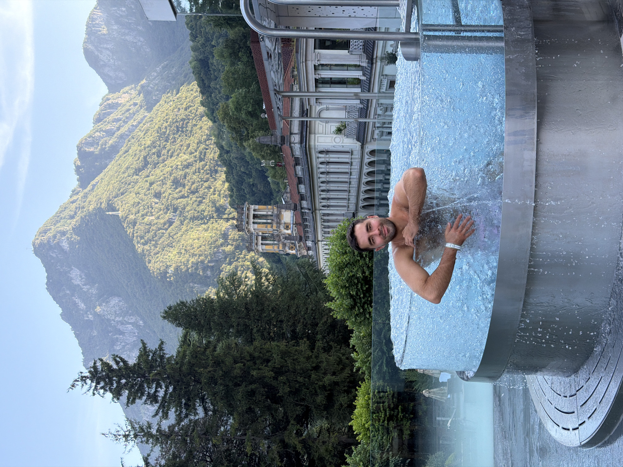 A man is partially submerged in a circular, bubbling hot tub with a transparent glass exterior, set against a backdrop of lush green mountains and a grand, classical building. The man, with dark hair and stubble, is looking towards the camera with a slight smile. He is wearing swim trunks. The hot tub's metal base features a ribbed texture, and water cascades down its sides. The building behind is ornate, with arched windows and a yellow tower. Tall evergreen trees are visible to the left of the building, and smaller trees and bushes form a line in front of it. The sky is a clear, pale blue. This scene is located in San Pellegrino Terme, Italy, likely at the Grand Hotel San Pellegrino or the associated spa.