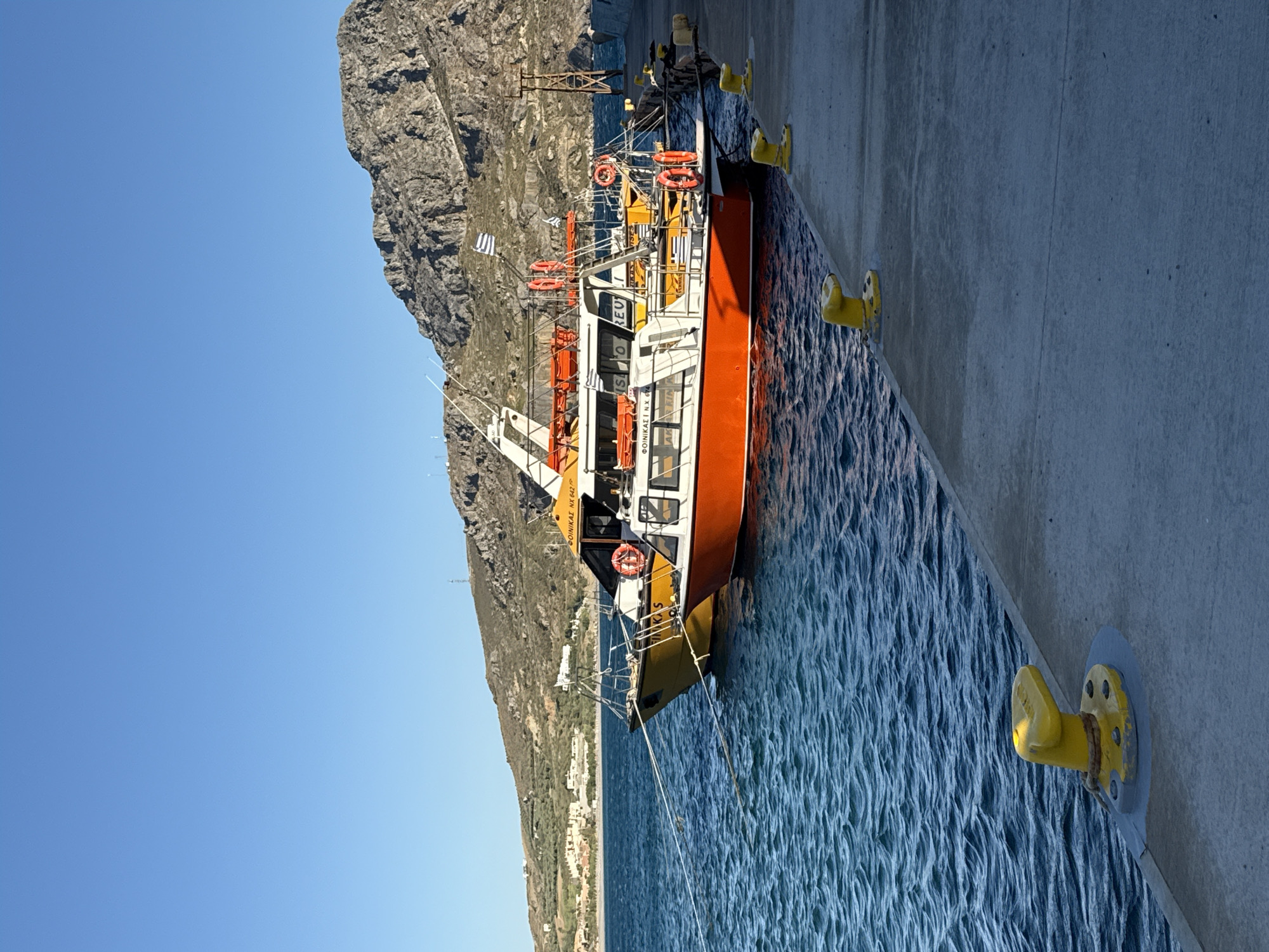 A brightly colored fishing boat with an orange hull and yellow trim is docked next to a concrete pier. The boat has a white cabin with windows and several life preservers are visible on its deck. The boat is tied to yellow cleats on the pier. In the background, a steep, rocky mountain rises behind the boat. The sky is clear and blue, suggesting a sunny day. The water is a deep blue and shows small waves. A town or village is visible on the hillside in the distance. The image appears to have been taken in the daytime, likely late morning or early afternoon. The overall scene is one of a peaceful harbor. Text on the boat reads "ΒΟΙΝΙΚΑΙ NX 642".
