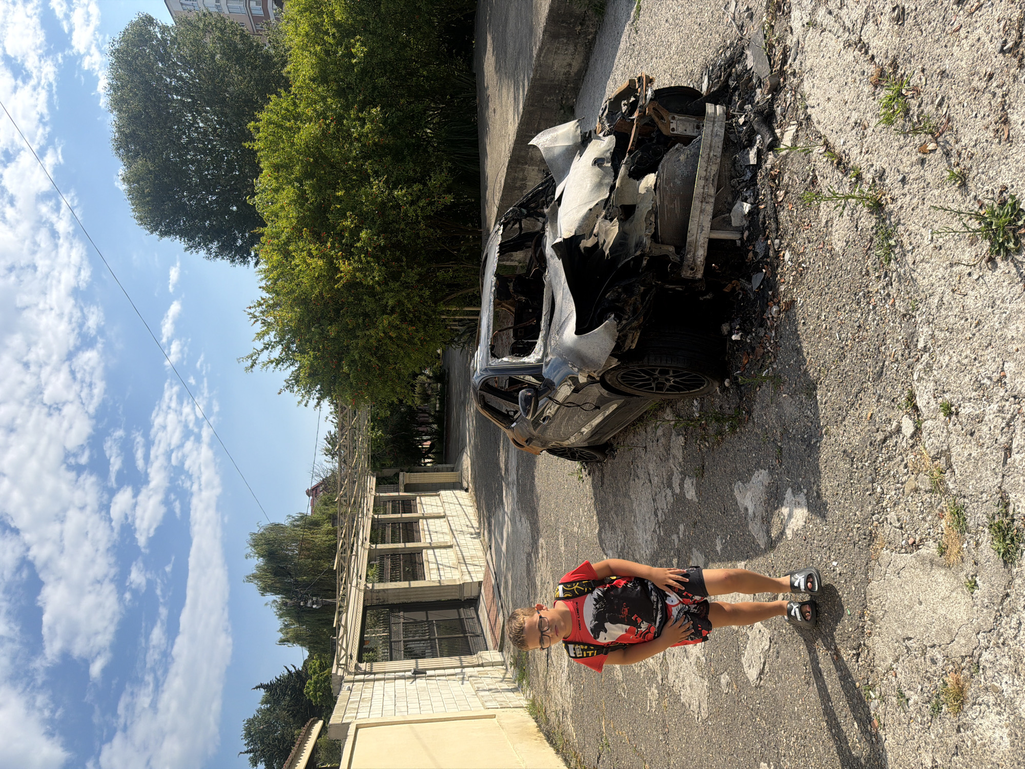 A young boy with glasses, wearing a red t-shirt with a black graphic and black shorts with red accents, stands on a cracked asphalt surface. He is looking towards the camera with a neutral expression. To his left, a heavily damaged and burnt car is parked on the side of the road. The car's interior and exterior show significant fire damage, with melted metal and charred remains visible. Behind the car and to the left of the boy, there is a partially constructed or dilapidated building with visible metal framework and windows. Green trees and foliage are present in the background, and the sky is blue with scattered white clouds, suggesting daytime with fair weather. A power line is visible stretching across the sky. The scene appears to be in an urban or semi-urban environment in Sochi, Russia, given the architecture and vegetation. There is no visible text in the image.