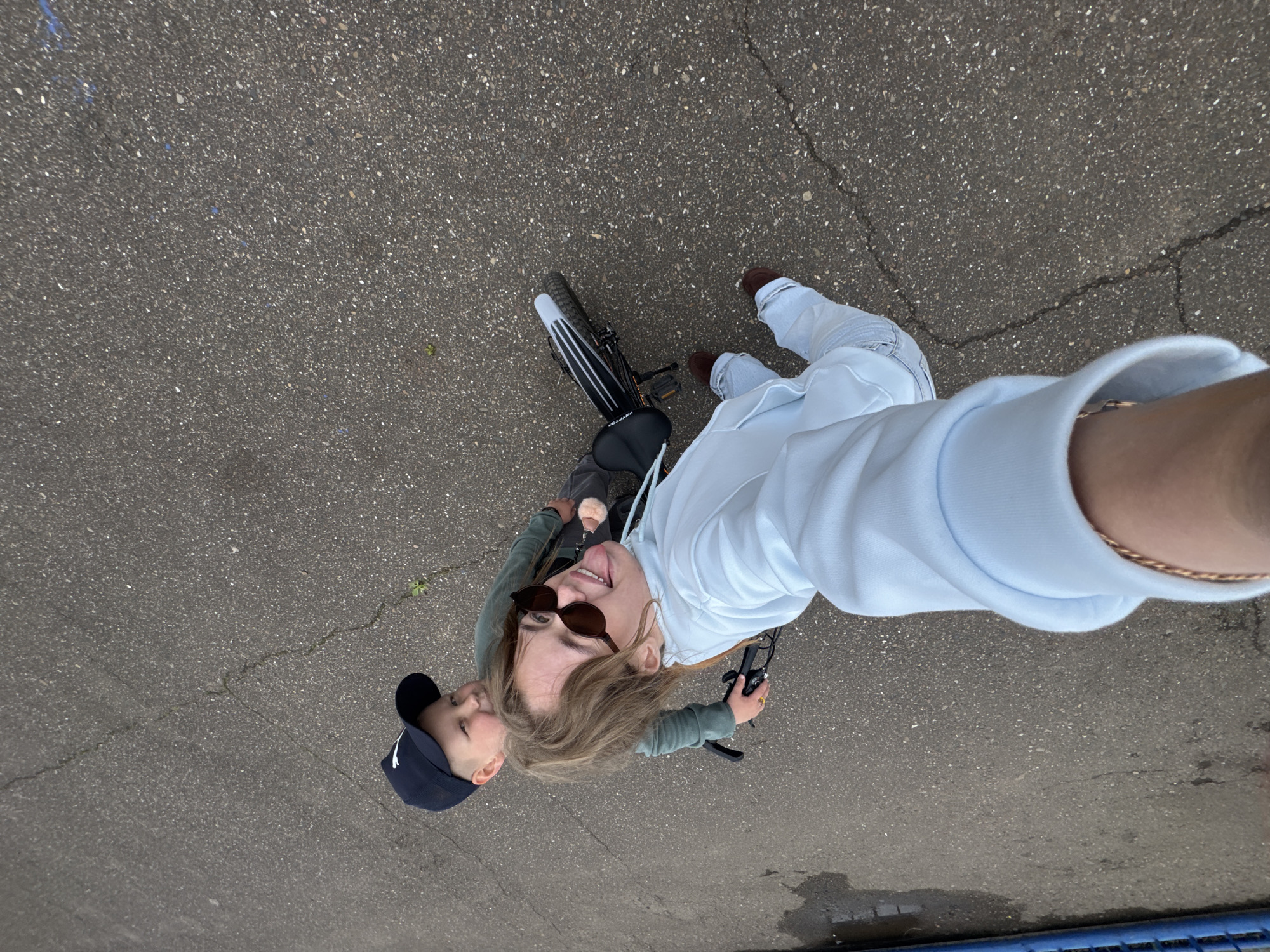 This is a high-angle, top-down outdoor image taken in the city of Chaikovsky, Russia, depicting two individuals on an asphalt surface.
In the foreground, a young adult or older youth, likely female, with long light brown hair and wearing dark sunglasses, a light blue hooded sweatshirt, light blue jeans, and dark brown shoes, is holding the camera at arm's length. She is playfully sticking out her tongue towards the camera. A gold-toned bracelet is visible on her right wrist.
Positioned behind and slightly to the left of her is a young child, likely male, wearing a dark blue baseball cap with a white logo (partially visible) and a dark green long-sleeved shirt. He is seated on a black bicycle, looking towards the camera with a subtle smile. The bicycle features a black seat with "VOLT" visible, a white rear fender, and thick tires. A small, light-colored pom-pom charm is attached near the first person's right shoulder.
The ground is dark gray asphalt pavement, characterized by visible cracks and scattered light-colored aggregate. A small green plant grows through one of the cracks. Wet patches or darker areas are present on the left side of the frame. A blue object, possibly a pipe or railing, is partially visible along the far left edge. The overall lighting suggests an overcast or diffused sky.