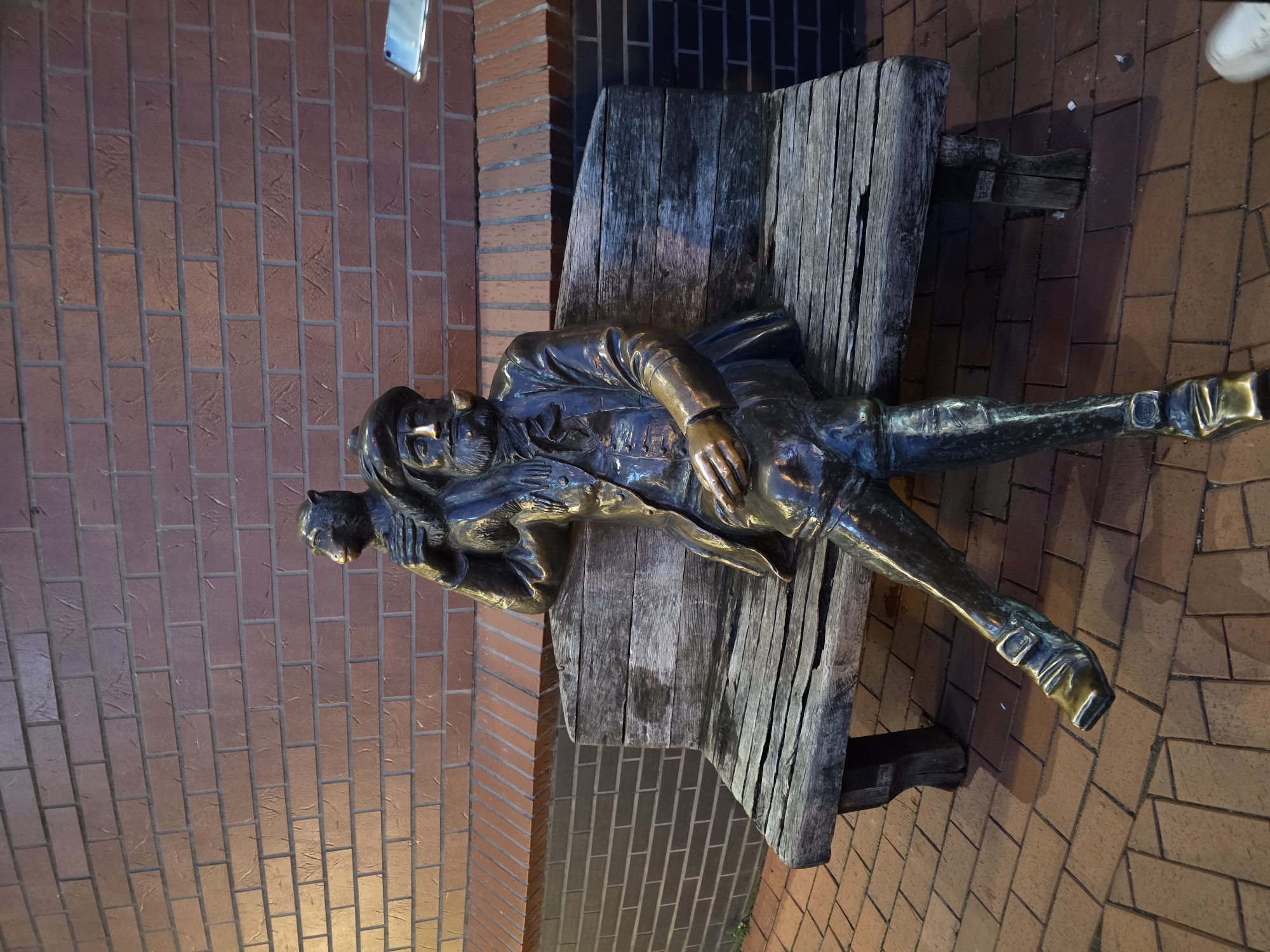 A bronze statue of a seated man with a pipe and a cat on his shoulder rests on a wooden bench. The statue is placed on a brick-paved sidewalk in front of a brick wall. The scene is lit by artificial lights, creating shadows and highlighting the textures of the materials. The statue's clothing appears to be from a historical period, with a hat, jacket, and breeches. The man has a beard and is looking to his left. The cat is perched on his left shoulder, facing the same direction as the man. The bench is weathered and shows signs of age. The brick wall behind the statue has a uniform pattern of red bricks. A portion of a smartphone is visible in the upper right corner of the image, reflecting the light. The overall impression is one of a public art installation. The statue is located in Kaliningrad, Russia.