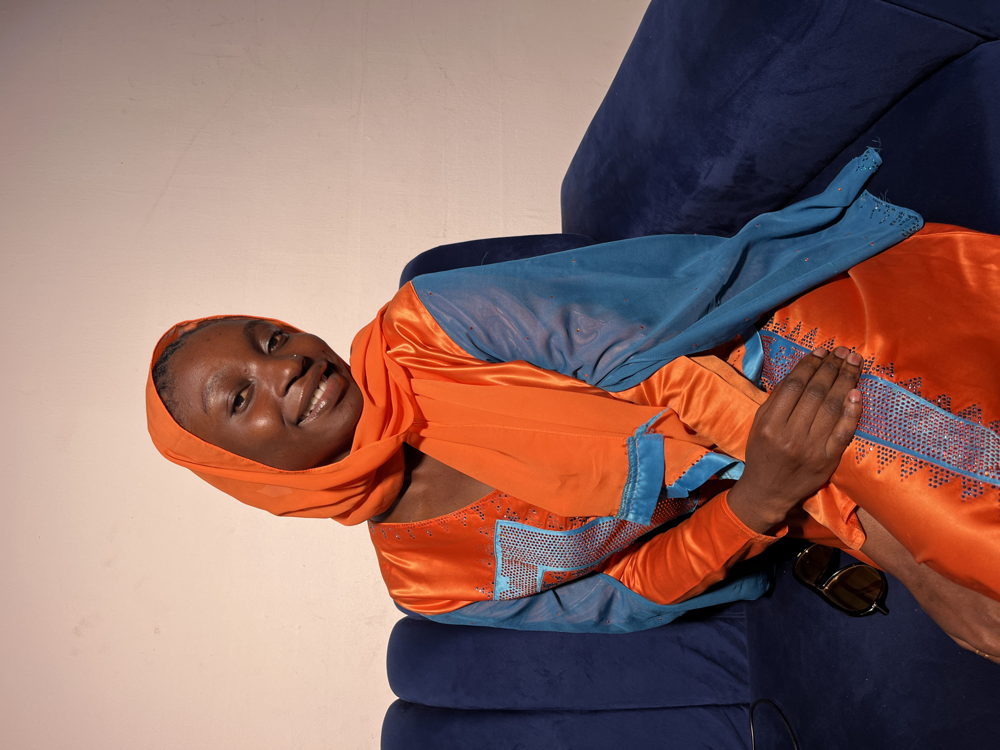 A young Black woman wearing an orange hijab and a shimmering orange and blue dress is sitting on a dark blue velvet couch. She is smiling at the camera. She has a nose ring and is wearing sunglasses on her lap. The background is a plain, light-colored wall.