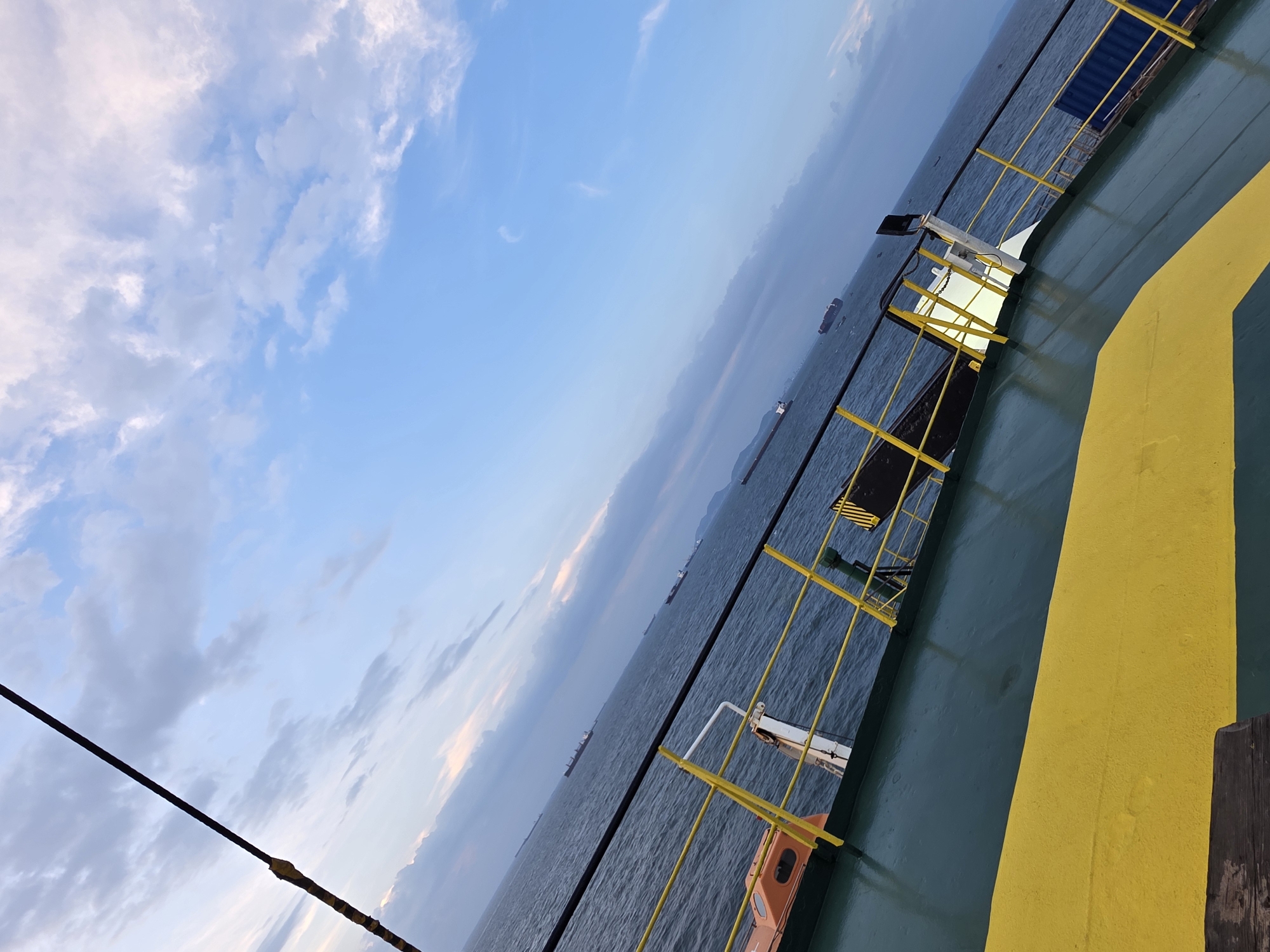 The image shows a view from the deck of a ship, looking out over the sea towards a hazy horizon. The sky is partly cloudy with shades of blue and gray. Several cargo ships are visible in the distance. The deck of the ship in the foreground is painted green with a yellow stripe. A yellow railing runs along the edge of the deck. An orange life raft is visible on the left side of the deck. The perspective of the image is tilted, suggesting it was taken from a moving vessel. The scene is likely in the waters off Vũng Tàu, Vietnam, a coastal city known for its shipping lanes and port activities.