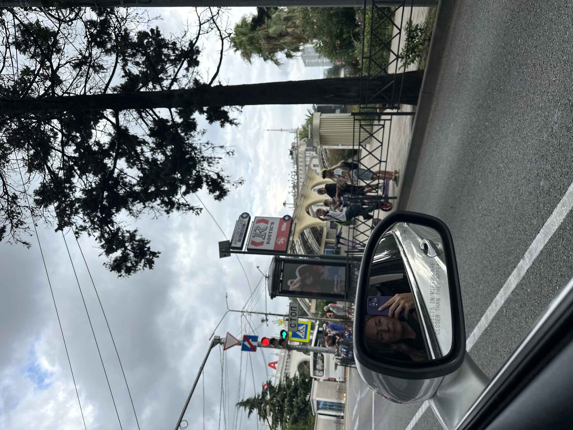 This image captures a street scene in Sochi, Russia, viewed from inside a vehicle. The foreground is dominated by a car's side mirror, reflecting a woman holding a purple smartphone to take a selfie. Her face is clearly visible, as is the ring on her finger. The mirror also shows the text "OBJECTS IN MIRROR ARE CLOSER THAN THEY APPEAR."
The background depicts a street with a traffic light showing a red signal, indicating a stop. Several people are visible on the sidewalk, some with electric scooters, suggesting a pedestrian area or a place where these are commonly used. A bus is parked on the side of the road. Behind the people, there's a building with an arched facade and advertising displays. A large tree with dark foliage partially obstructs the view of the sky, which is overcast. In the distance, urban buildings and greenery are visible. A sign with Cyrillic lettering, "АВТОБОКЗАЛ" (Autovokzal, meaning Bus Station), is prominently displayed.