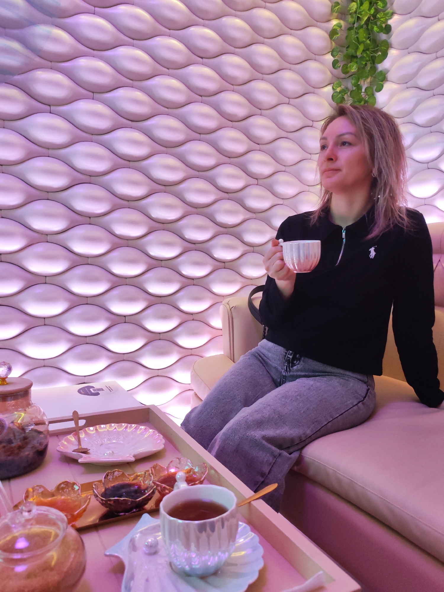 A woman is seated on a pale pink sofa in what appears to be a lounge or spa setting. She wears a black, long-sleeved polo-style shirt with a white logo on the chest, and gray jeans. She holds a small white cup in her right hand, near her face, and appears to be looking off to the side.
A low, light-colored tray table is positioned in front of her. On the tray, there is a variety of items, including a teapot, several small dishes containing what appear to be jams or sauces, a white shell-shaped dish, and a teacup on a saucer. A small wooden spoon rests on one of the dishes.
Behind the woman is a decorative wall featuring a repetitive, rounded wave pattern with a light-pink hue. A few green vines are visible in the upper right corner of the frame, near the top of the wall. The lighting in the scene is soft and diffused, creating a calm and relaxing atmosphere.
The location is identified as Izhevsk, Russia.