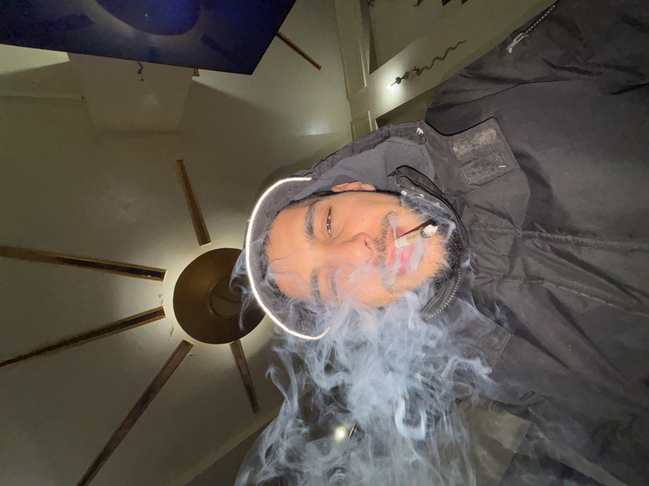 An adult male with a beard and mustache is depicted in a close-up, upward-angled shot within an indoor setting in Tanger, Morocco. He wears a black hooded jacket, the hood of which features a prominent white or reflective trim, forming a semi-circular halo effect above his head. A lit cigarette is held between his lips, and a large volume of white-grey smoke billows from his mouth and around his face, obscuring the left side of his head. His eyes are open, and he appears to be looking directly forward. The background reveals a light-colored ceiling with a central circular light fixture from which eight dark-colored, radiating beams extend, forming a sunburst or fan pattern. To the upper right, a dark, rectangular object, possibly a screen or artwork, is mounted on the wall. The scene is illuminated by artificial light, primarily from the ceiling fixture. No specific street or landmark within Tanger is discernible.