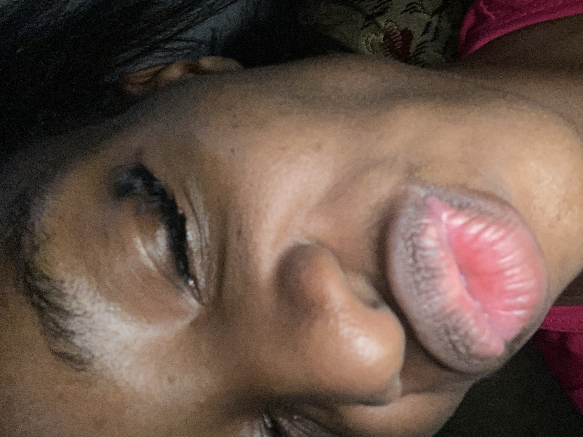 The image is a close-up, low-angle shot of a person's face, focusing on their lips and nose, with one eye partially visible. The person appears to be making a kissing gesture. The skin tone is dark, and the lips are pink and plump with visible texture. The background is dark and out of focus, with a suggestion of patterned fabric on the right side. No specific location in the city of Jos, Nigeria can be determined from this image.
