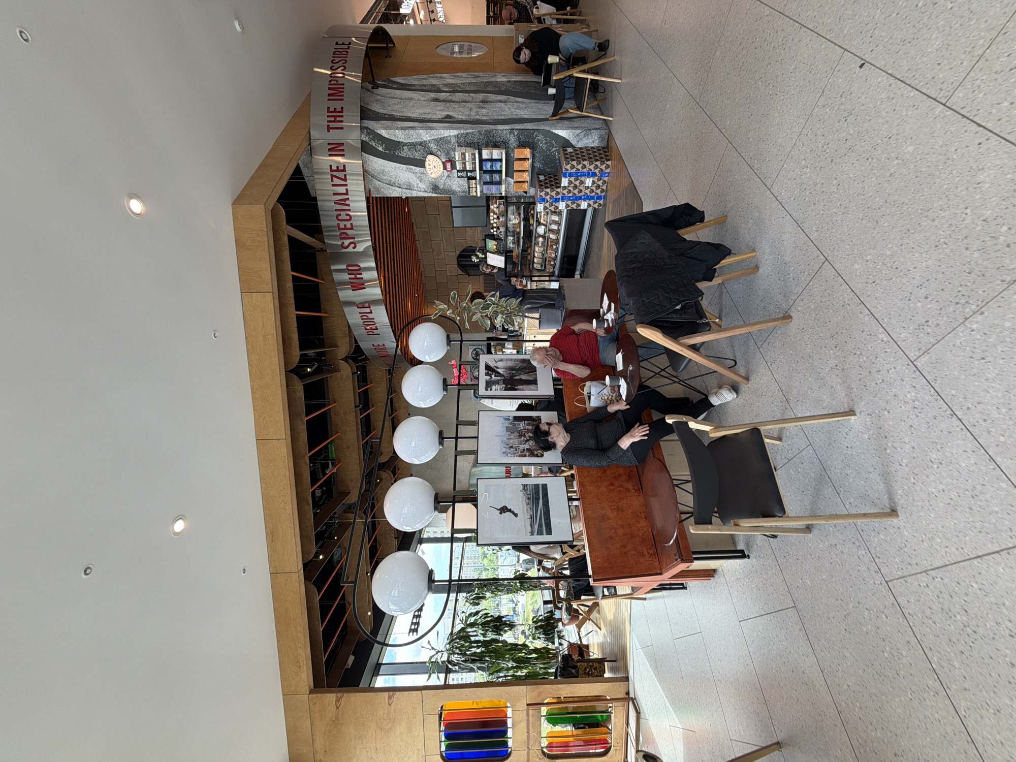 This image shows the interior of a cafe or restaurant. The location appears to be a bright, modern space with a neutral color scheme. There are people seated at tables, apparently relaxing or conversing. One person is wearing a red shirt, while another is wearing black. The furniture includes wooden chairs and tables. There is a serving counter with a display of food items. The environment includes decorative elements such as hanging spherical lights, framed photographs, and plants. The signage includes the phrase "MORE PEOPLE WHO SPECIALIZE IN THE IMPOSSIBLE" in red lettering. Colorful glass bricks can be seen near the bottom. The city and state are unknown.