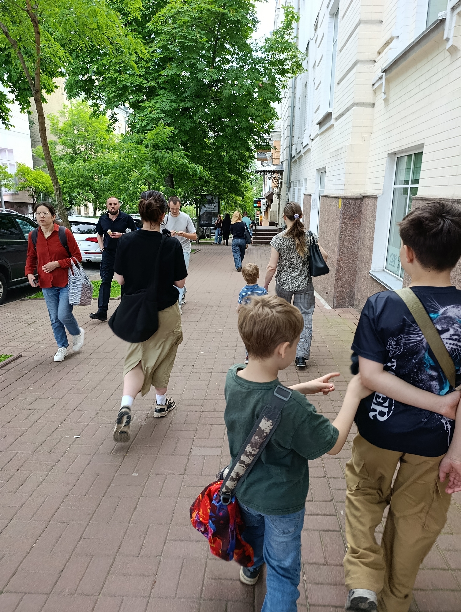The image depicts a group of people walking on a brick sidewalk in Kyiv, Ukraine. The environment includes a light-colored brick building on the right and lush green trees on the left, suggesting a city street with foliage. Several individuals are visible, including adults and children.
In the foreground, two young boys are walking away from the camera. The boy on the left wears a dark green t-shirt and blue jeans, carrying a bag with a colorful pattern. The boy on the right is seen from the back, wearing a dark t-shirt with a graphic print and khaki pants, with an arm around the shoulder of the boy in front.
Further ahead, a woman in a black t-shirt and khaki shorts walks away from the camera, carrying a large black bag over her shoulder. Beside her, a man in a light-colored t-shirt and dark pants also walks away. To the left, a woman in a red shirt and blue jeans walks towards the camera, carrying a striped bag and wearing glasses. Behind her, a man in a black t-shirt and dark pants is visible, and a car is parked on the left side of the street.
Another group further down the sidewalk includes a woman in a patterned shirt and grey pants, and a young child in a blue and white striped shirt walking ahead of her. The overall scene captures a moment of daily life on a street in Kyiv.