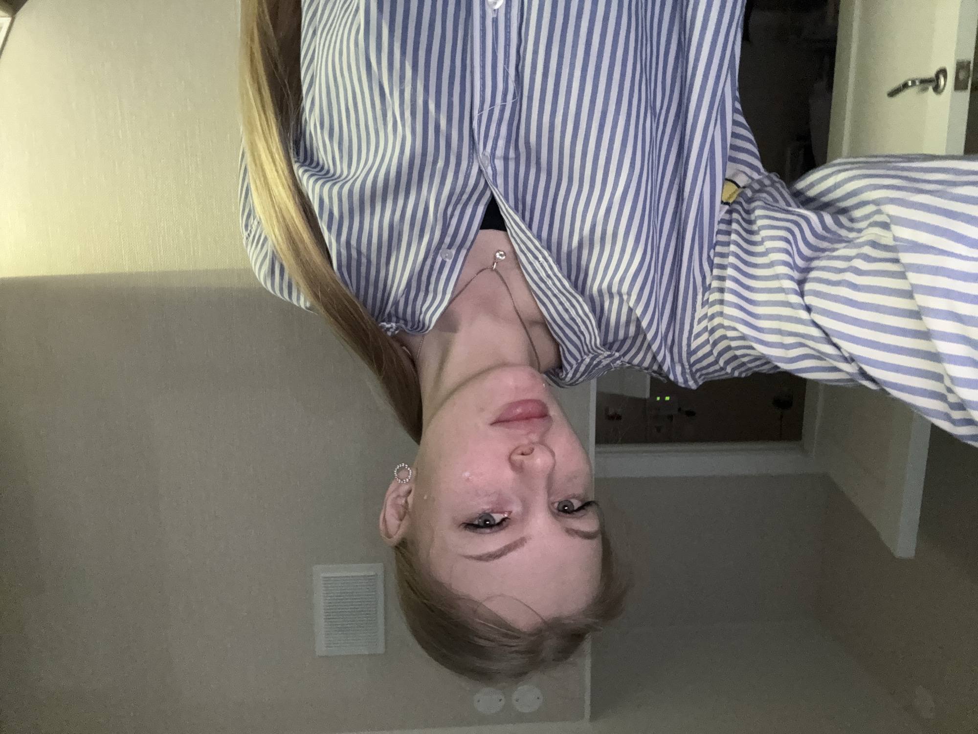 A photograph, likely taken indoors, presents a person from an upside-down perspective. The subject appears to be a young woman with fair skin and blonde hair. She is wearing a blue and white striped, button-down shirt, and a necklace is visible. Her earrings are small and circular. The background shows a cream-colored wall with a square ventilation grill. Part of a doorway is visible in the background. Based on available information, there is insufficient data in the provided image to determine if the photo was taken in Perm, Russia.