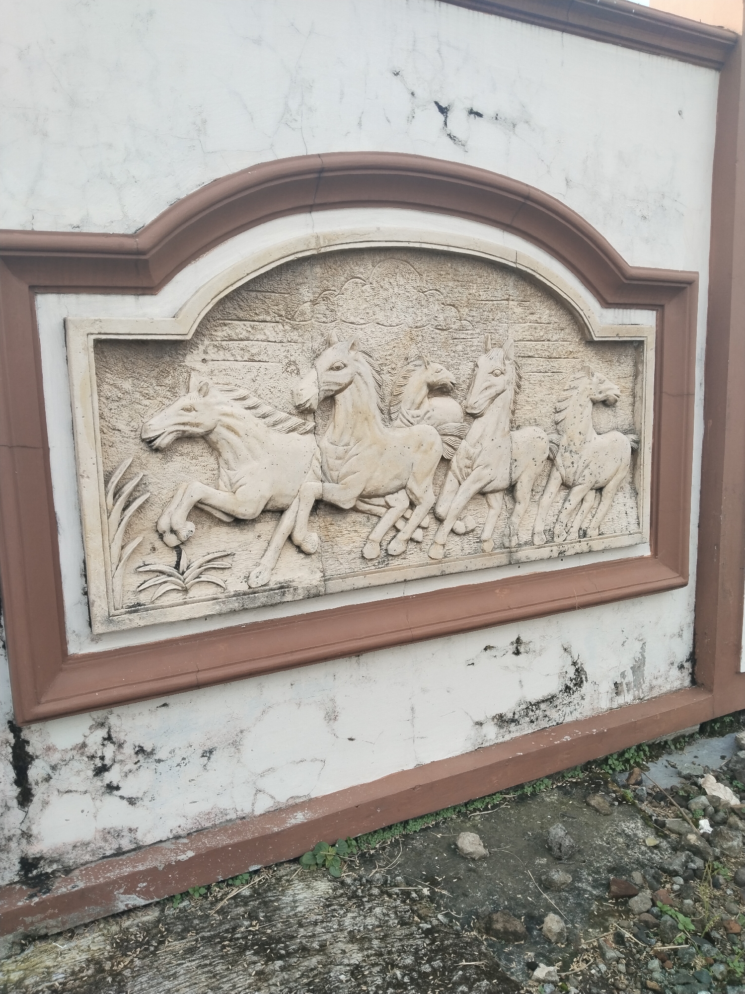 An outdoor scene in Bangsri, Indonesia, features a relief sculpture of horses embedded in a white wall. The sculpture, carved in a light, off-white material, depicts six horses in various dynamic poses, appearing to be running or galloping from left to right. The lead horse on the far left rears slightly, while the others follow closely behind, conveying a sense of movement and energy. Stylized plant motifs are visible on the left side of the relief, near the lead horse's front legs, and the background of the carving has a textured, rough appearance.
The relief is framed by a decorative border, which is light beige on the inside and a prominent reddish-brown on the outside. This framed section is set into a larger white wall, which shows significant signs of age and exposure to the elements, including numerous cracks, dark smudges, and patches of dirt or mold. The top of the wall is also bordered by a matching reddish-brown trim.
At the bottom of the image, the ground transitions from concrete to a patch of dirt and loose rocks, with some small green weeds growing sporadically. The lighting suggests it is daytime, with no shadows indicating a specific time, and the weather appears clear. There is no visible text in the image.
