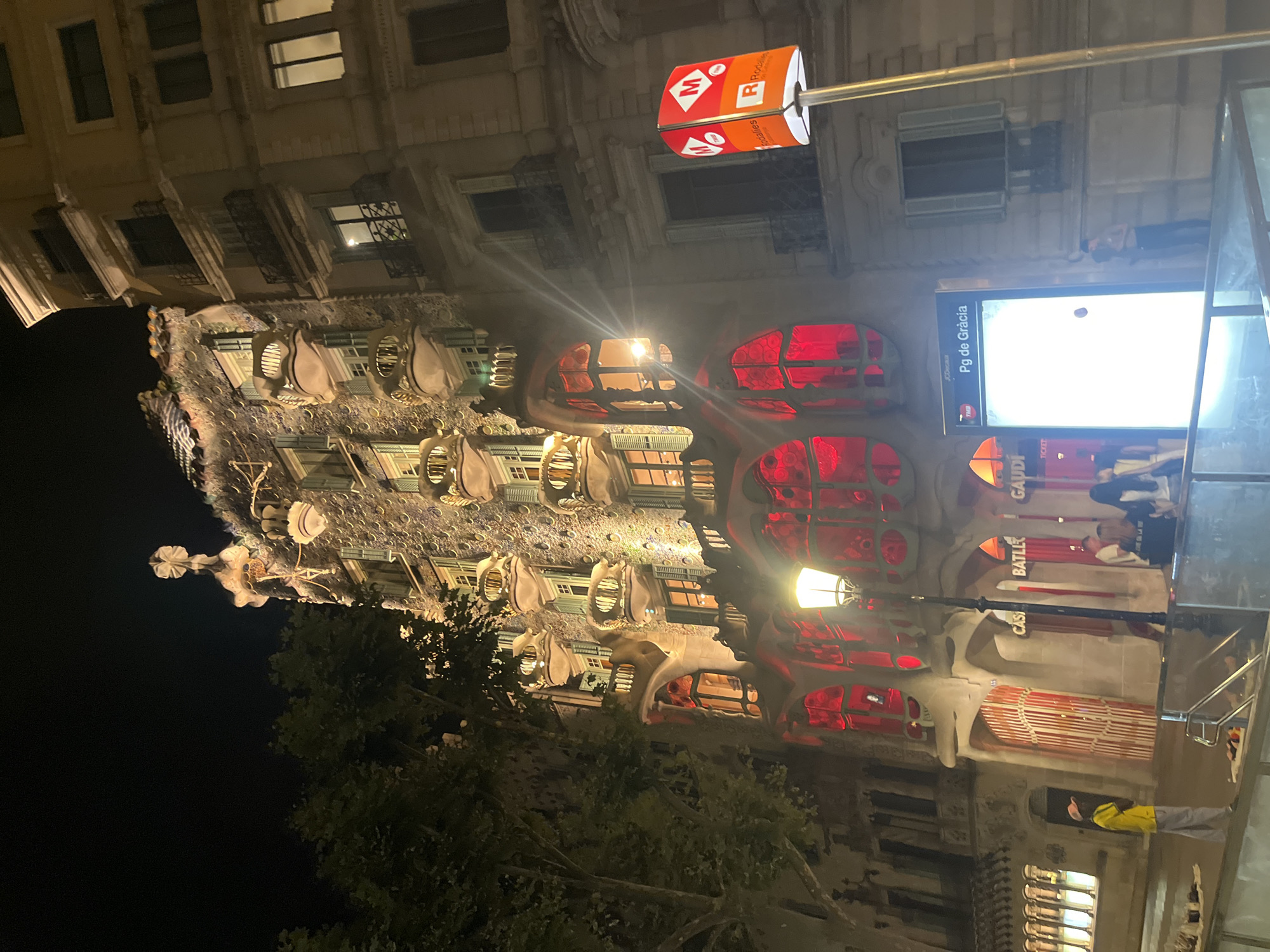 The image captures a nighttime view of Casa Batlló, a modernist building designed by Antoni Gaudí, located on Passeig de Gràcia in Barcelona, Spain. The facade of Casa Batlló is illuminated, highlighting its distinctive organic shapes, balconies resembling skulls, and a roof that resembles the scales of a dragon. Red lights emanate from the large, rounded windows on the lower floors.
To the right of Casa Batlló, a neighboring building with a more traditional architectural style stands, its windows also lit up. In the foreground, a street lamp with a warm glow is visible. A public transportation sign for "Pg de Gràcia" with the "M" logo for metro services is also present.
Several people are visible. A person wearing a yellow shirt and grey pants stands near the entrance of Casa Batlló, facing away from the camera. Other individuals are seen in the background, some standing and others appearing to be entering or exiting the building. The street is paved, and the overall environment suggests an urban setting at night.