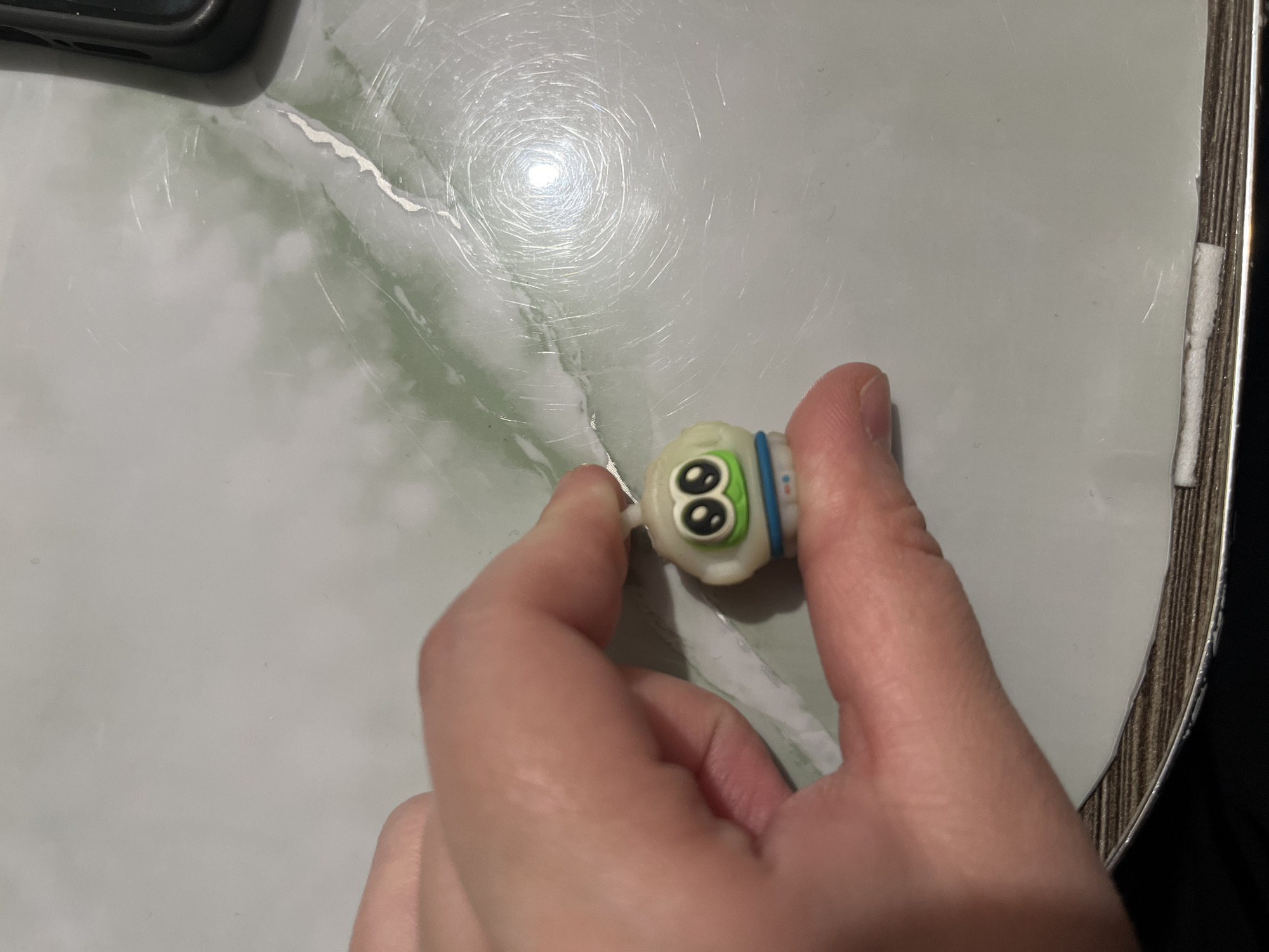 A person's hand is holding a small, off-white toy with large, cartoonish eyes and a blue band around its neck. The toy is held against a background of a marble-patterned surface, likely a table. A portion of a dark-colored object, possibly a phone, is visible in the upper left corner of the frame. The setting appears to be indoors, and the location is unknown.