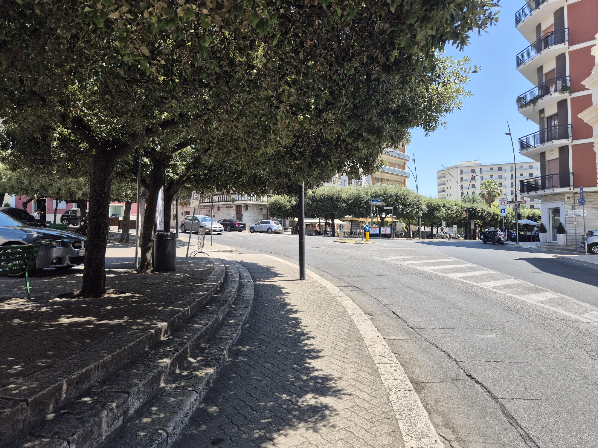 The image captures a sunlit street scene in Laterza, Italy, specifically on Via Pietro Nenni. The perspective is from a raised sidewalk with stone steps leading down towards a street that curves to the right. Lush green trees with dappled shadows cast by the sunlight line the left side of the street, creating a shaded pedestrian area. Several cars are parked along the sidewalk and a few are driving on the street. Buildings, including a modern apartment building with balconies, are visible in the background. The street itself is paved with asphalt and marked with white lines. In the distance, a roundabout with traffic lights and small trees can be seen.