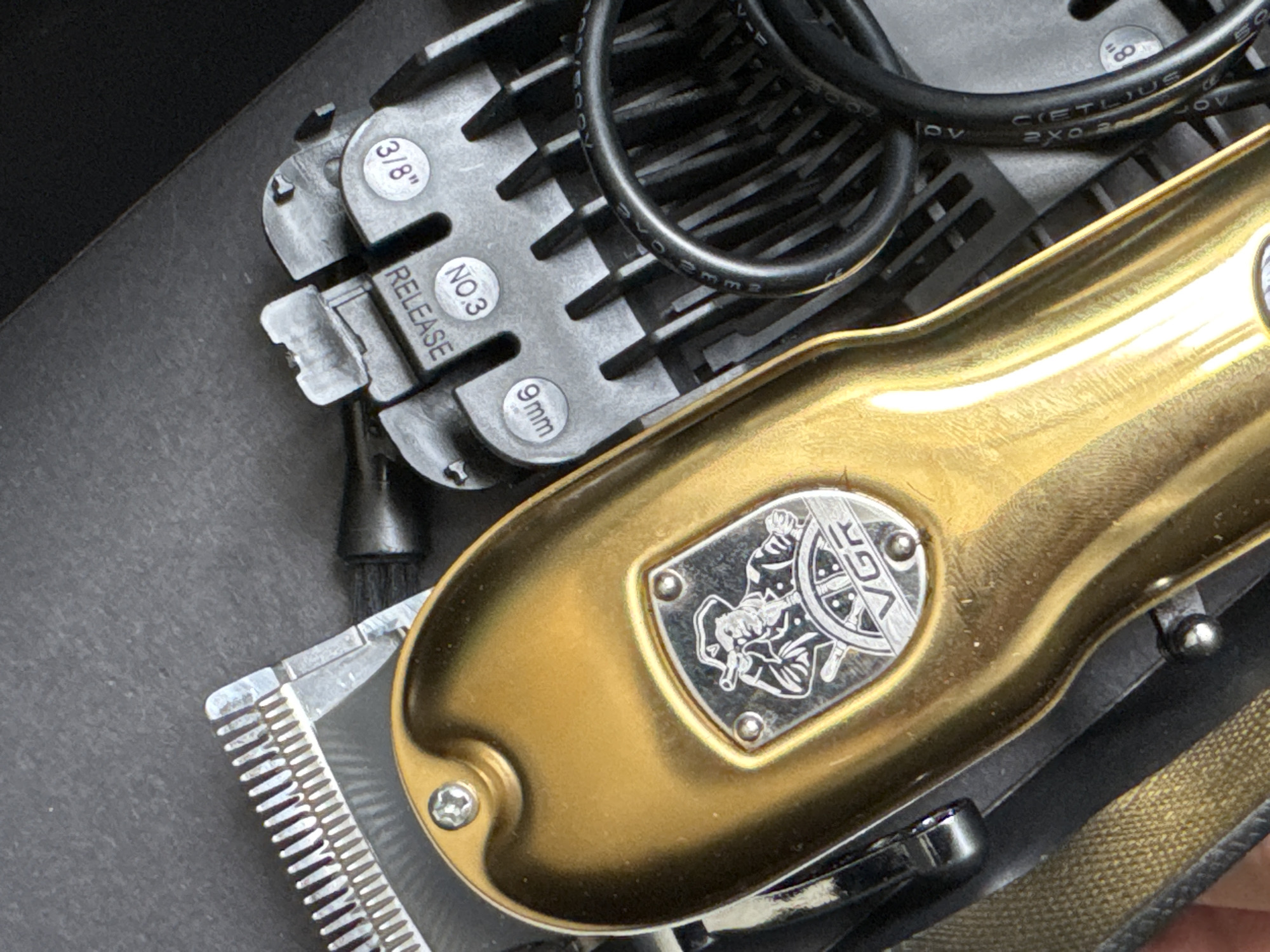 The image shows a close-up, high-angle shot of a set of hair clippers and accessories, resting on a dark surface. The main subject is a gold-colored clipper with a silver emblem featuring a man in a pirate hat holding a spyglass and steering a ship's wheel. The brand name "VGR" is visible on the emblem. To the right of the clipper, there is a black plastic attachment with several comb guards. Some of these guards are labeled with numbers and measurements, including "NO. 3", "9mm", and "3/8"". A black power cord is coiled around the attachment. In the top left corner, part of a silver clipper blade with fine teeth is visible. The background is a dark, textured surface, possibly fabric or a dark box. The scene is well-lit, with reflections on the gold clipper indicating the presence of a light source. The location in the city of Яромінскі сельскі Савет, Belarus is not determinable from the image.