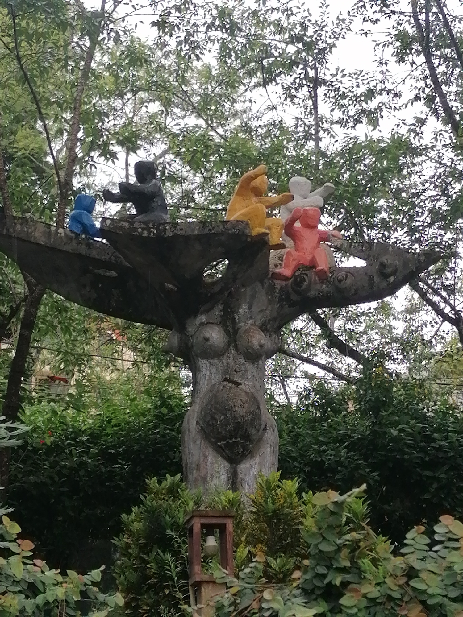 The image shows a concrete sculpture in a garden setting in Da Lat, Vietnam. The sculpture is of a large, stylized tree trunk with prominent breasts, resembling a female torso. Several smaller figures, appearing to be children, are perched on the branches and trunk. One figure, in blue, is sitting on the left side. To its right, a dark-colored figure is seated. Further right, a yellow figure is seated, and next to it, a white figure is standing. On the far right, a red figure is seated. The sculpture is surrounded by lush green foliage and trees. In the foreground, there is a small, wooden structure with a light fixture. The overall impression is of a whimsical and somewhat abstract art installation in a natural environment.