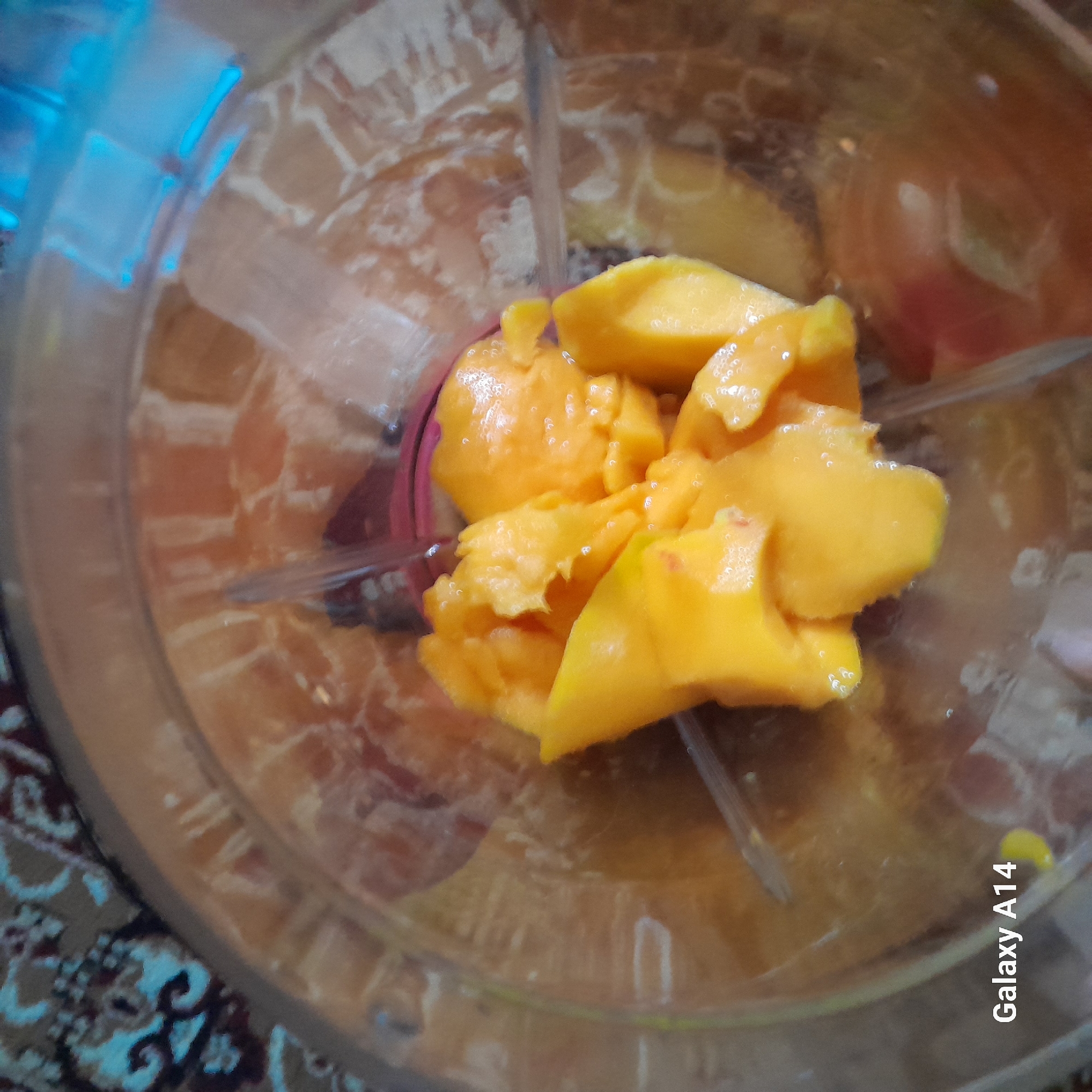 The image shows a close-up, overhead view of cut pieces of mango inside a blender pitcher. The mango pieces are a vibrant orange-yellow color and appear to be fresh and juicy. The blender blades are visible in the center, surrounded by the mango. The background is a blurred pattern of a carpet or textile. The lighting in the image is bright, causing some glare and highlights on the mango and the blender. The bottom left corner of the image has the text "Galaxy A14" watermark. The image is a still photograph, with no indication of movement or action.