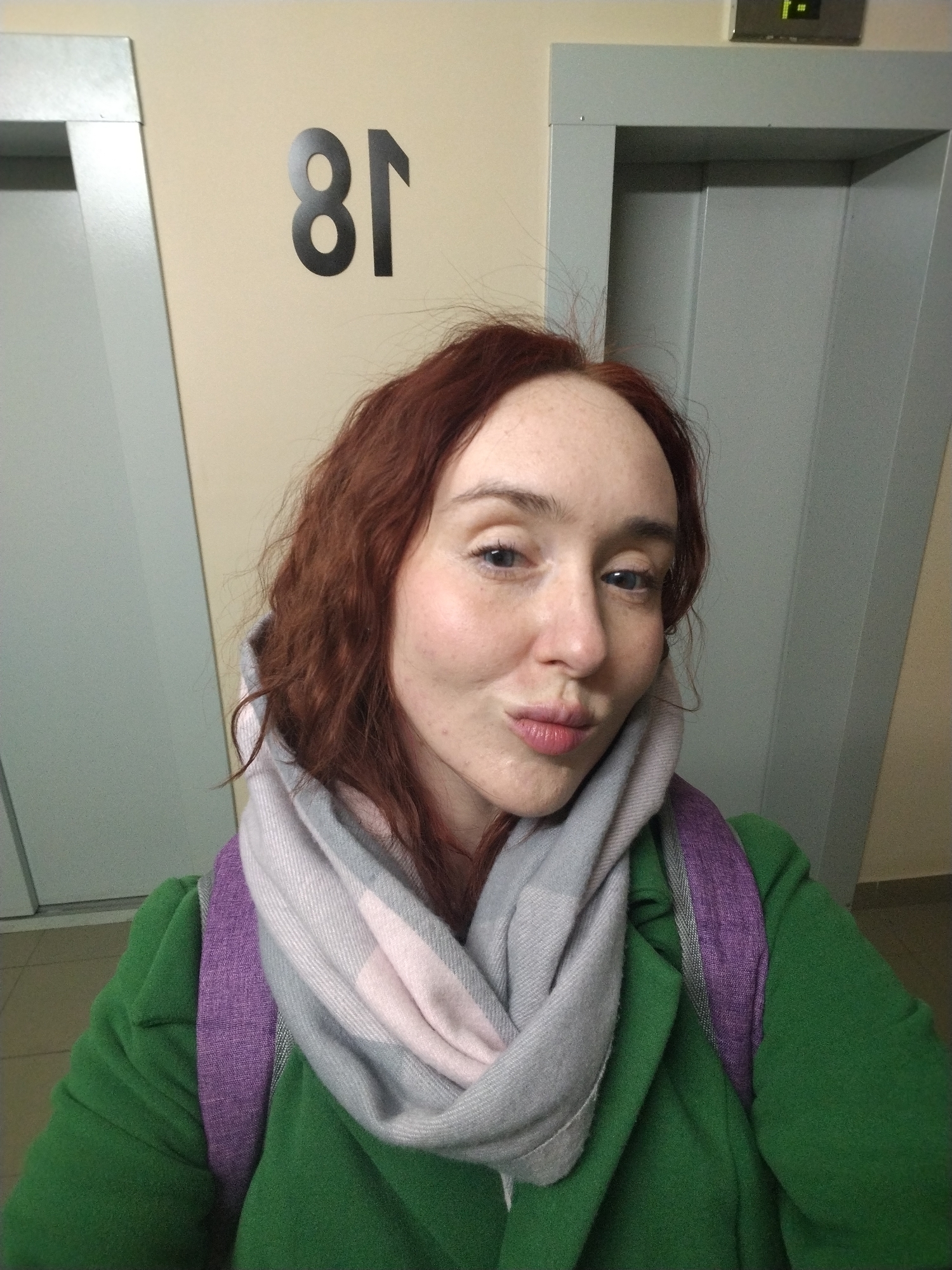 A woman with red hair and freckles is taking a selfie in what appears to be an elevator lobby. She is wearing a green coat, a gray and pink scarf, and a purple backpack. Behind her are two elevator doors and a sign that reads "18" in reverse, indicating that it is likely in Russia. Based on the architectural style and the elevator sign design, the location is possibly in a residential building in Izhevsk, Russia. The woman is making a kissing face at the camera.
