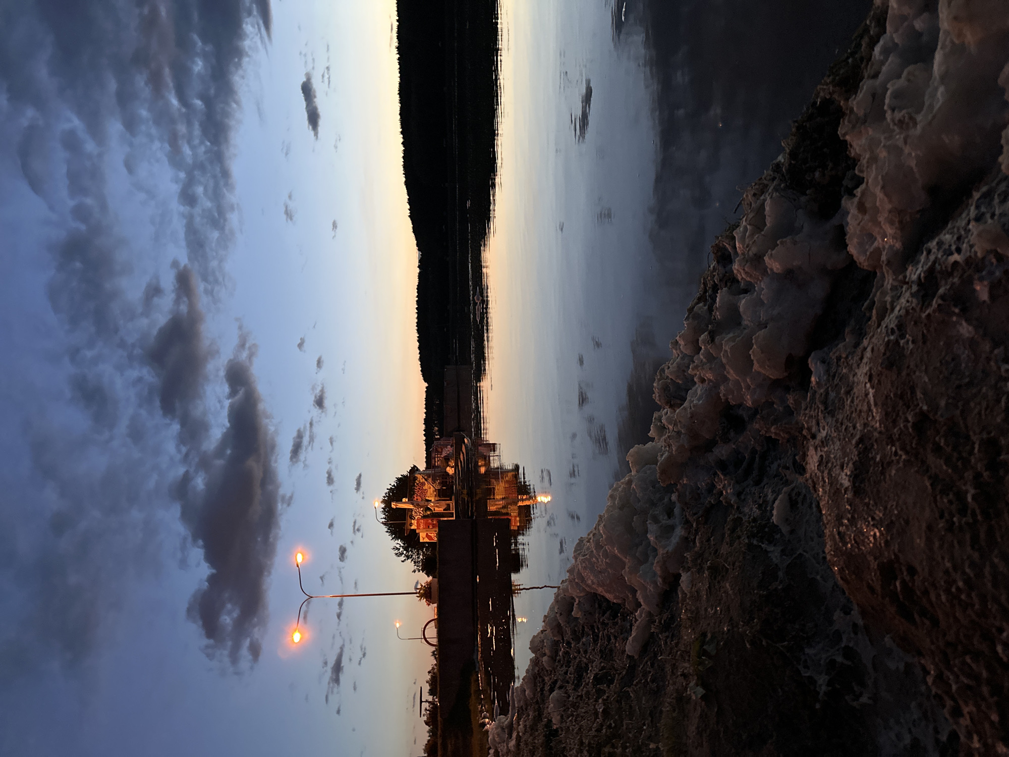 The image depicts a waterfront scene at dusk, likely in Minsk, Belarus, given the presence of a Soviet-era style lamppost and what appears to be a food kiosk reminiscent of those found in Eastern European cities. The sky is a deep blue with dramatic clouds illuminated by the fading sunlight, creating a soft gradient towards the horizon. In the foreground, the edge of the water is lined with a frothy, white substance, possibly foam or algae buildup, with a rocky, uneven texture.
Dominating the mid-ground is a concrete embankment where a double-headed lamppost stands, casting a warm, orange glow. Adjacent to it, a small, multi-story food kiosk or building with lights on is visible, its reflection shimmering on the still water. The kiosk appears to have signage, including what looks like a Coca-Cola logo, and is adorned with lights. A tree stands behind the kiosk, its silhouette dark against the sky. A bicycle rack is also present on the embankment.
The water itself is calm and reflective, mirroring the sky, clouds, and the lights from the kiosk. The opposite bank of the water body is lined with a dense forest, appearing as a dark, silhouetted band against the twilight sky. The overall atmosphere is serene and slightly melancholic, characteristic of the transition from day to night. There are no people visible in the image.