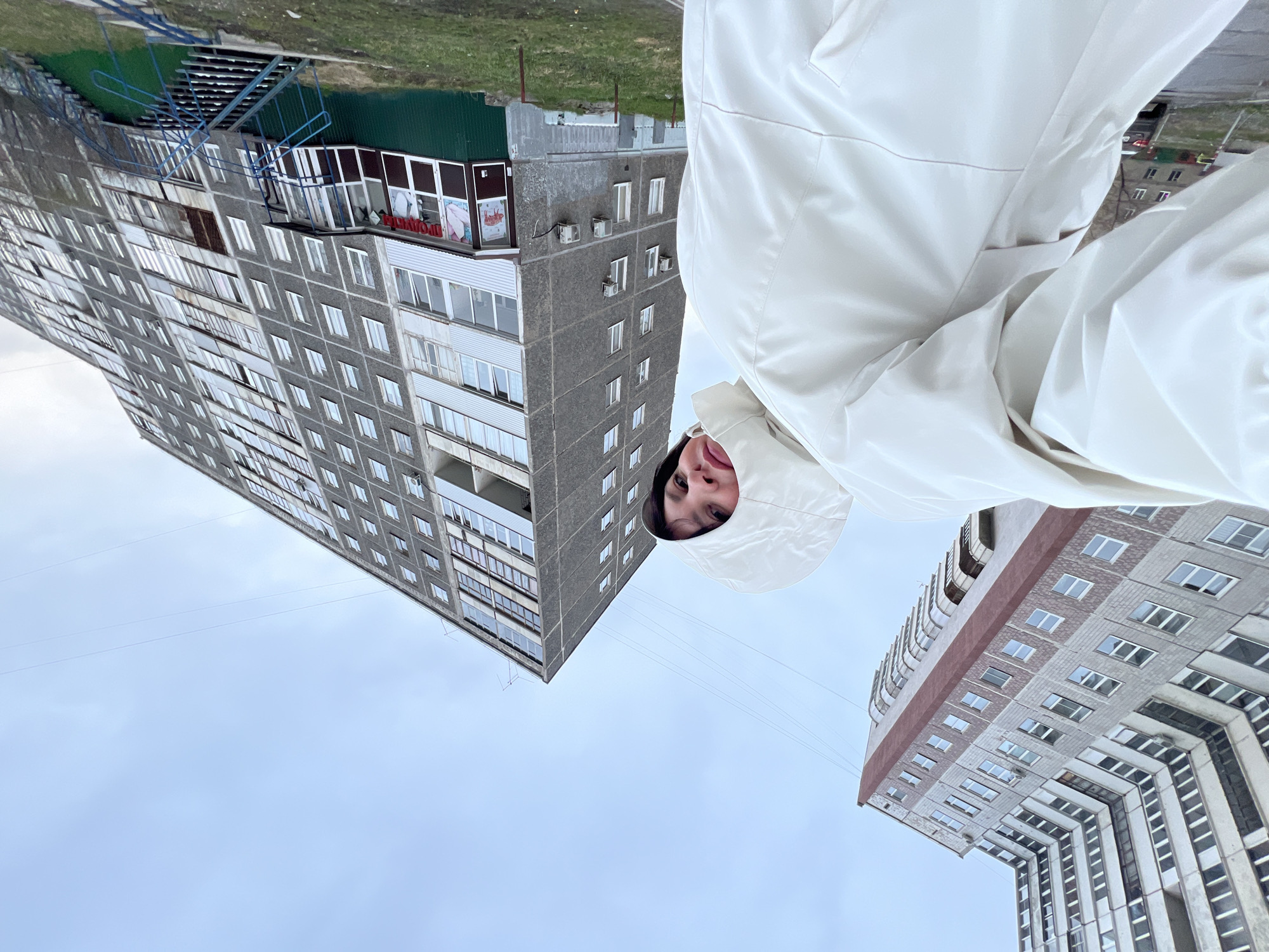 This low-angle photograph captures a person looking upwards in an urban setting, likely Perm, Russia. In the immediate foreground, a young person, likely female, is partially visible, framed by the hood of a light-colored (white or cream) jacket. Only her face, including her eyes, nose, and mouth, is seen looking directly at the camera with a neutral or slightly smiling expression.
The background is dominated by two large, grey, multi-story concrete apartment buildings, characteristic of Soviet-era architecture. The prominent building on the left features numerous windows and balconies across its facade. At its base, there's a commercial unit with large glass windows, a dark green corrugated metal roof, and an external blue metal staircase. A red sign on this unit partially displays the Russian word "ПРОДУКТЫ" (PRODUKTY), meaning "Groceries" or "Products." Another tall apartment building is visible on the right, partly obscured by the person's jacket, also featuring multiple windows and a reddish-brown band along one section.
The sky is overcast and light grey, suggesting a cloudy day with soft, diffused light. Several thin wires stretch across the sky between the buildings. A patch of green ground or grass is visible above the left building, further emphasizing the upward perspective of the shot. The person's hooded jacket suggests cool or cold weather conditions.
